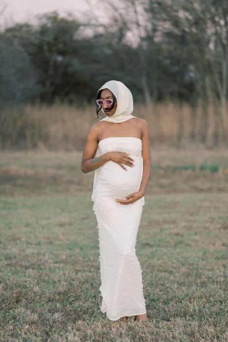 A person draped in white fabric stands in a grassy field during a soft-lit photoshoot.