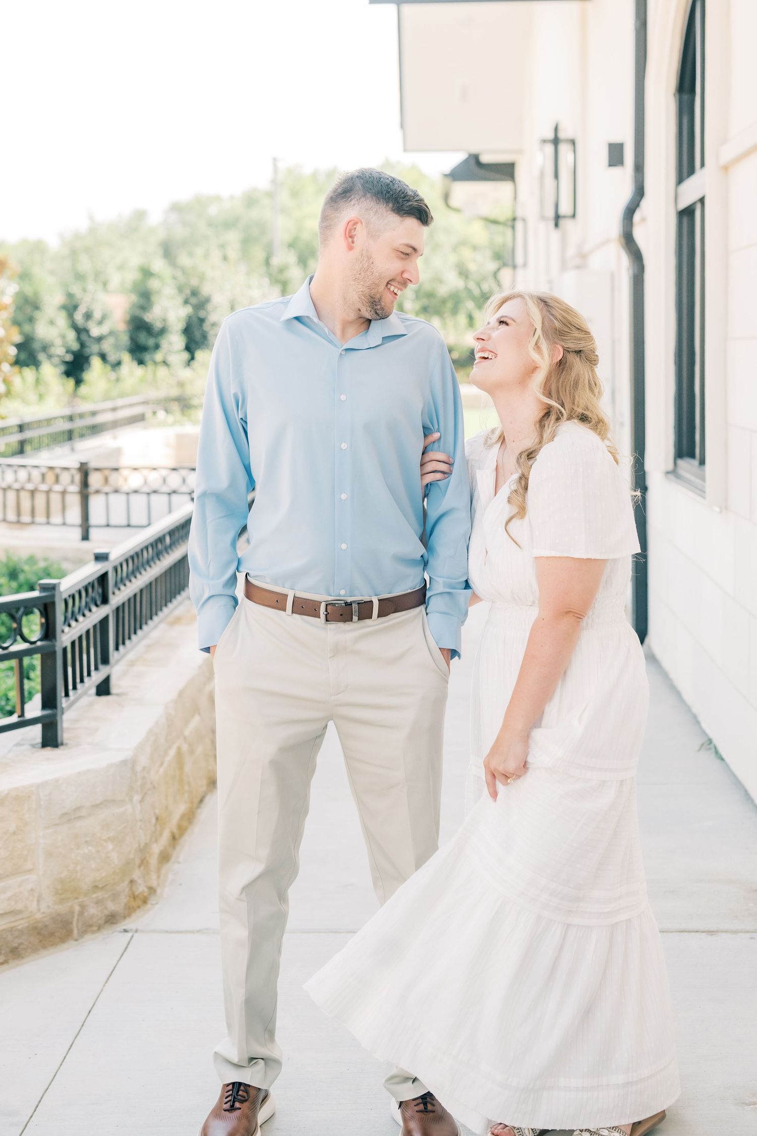 Happy couple laughing together outdoors on a sunny patio, man in blue shirt and woman in white dress.