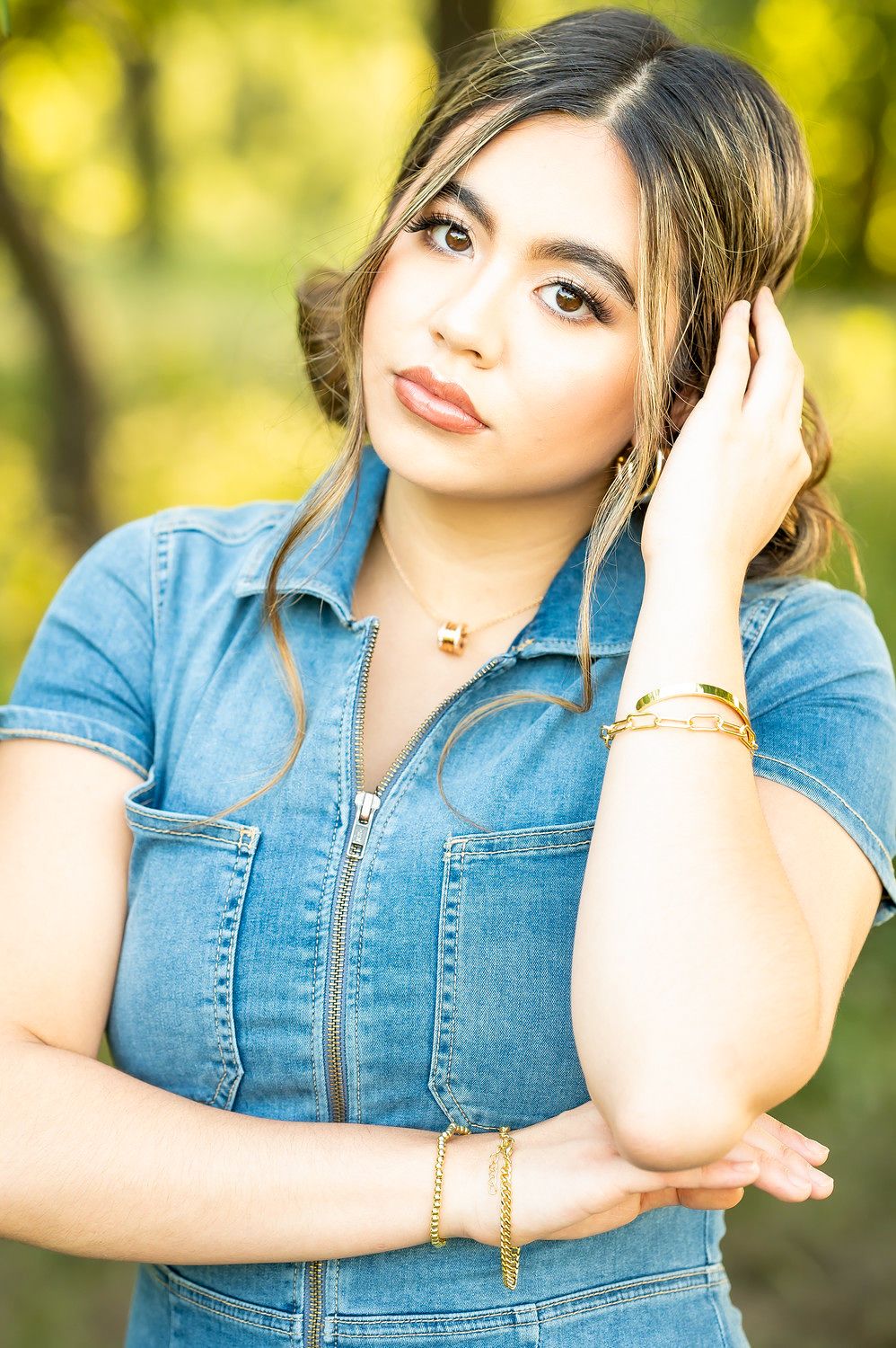 Young woman in denim jumpsuit posing outdoors with gold jewelry in natural light.