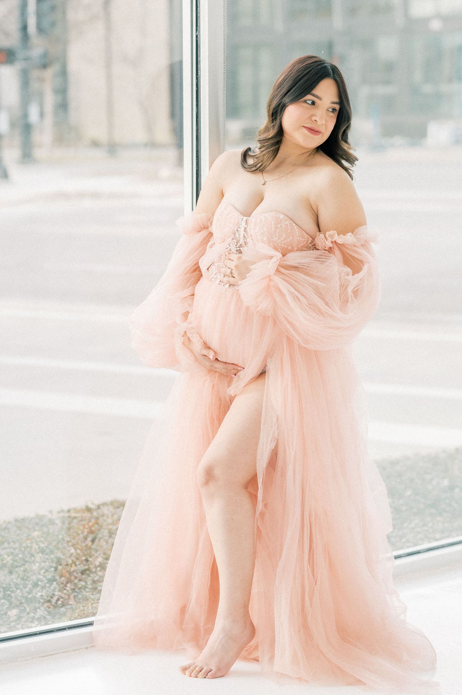 Pregnant woman in a flowing blush pink tulle maternity gown poses barefoot by a large window.