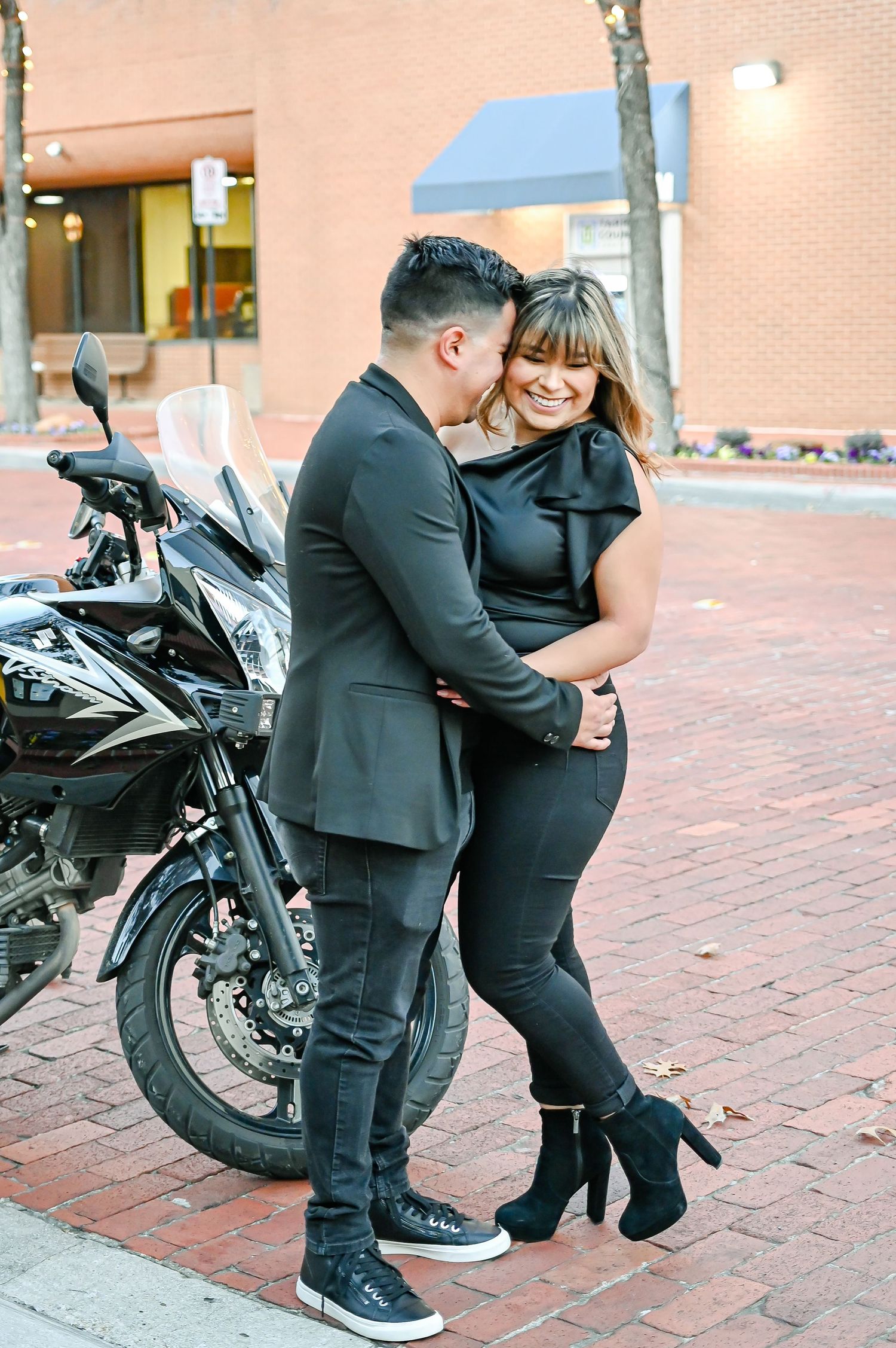 Romantic couple embracing and laughing together beside a black motorcycle on a brick street.