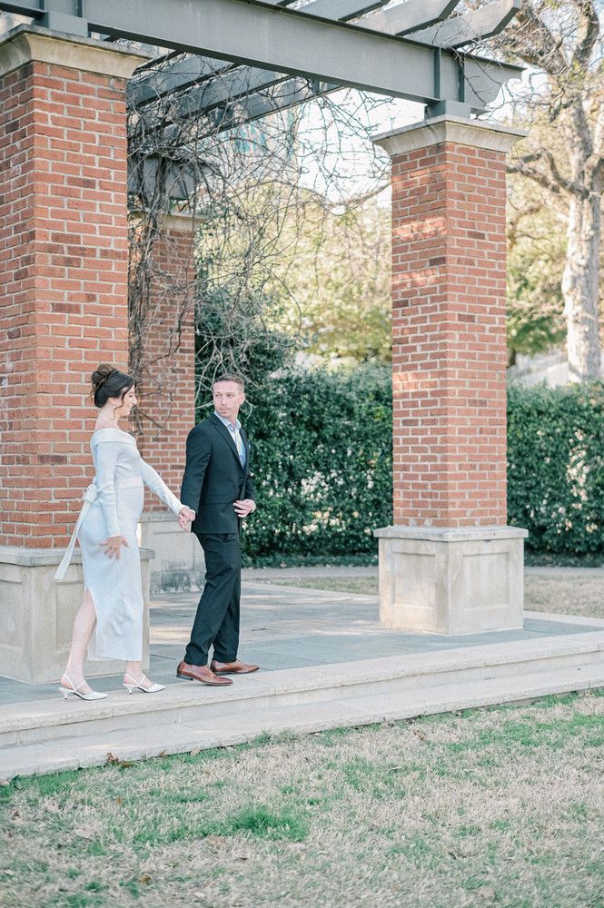Bride and groom holding hands walking under brick pergola in outdoor garden setting on wedding day.