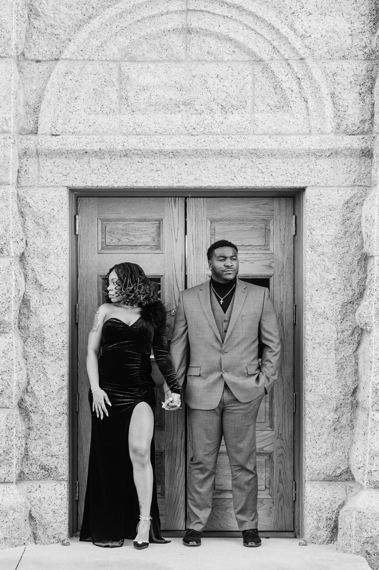 Stylish couple holding hands before grand wooden doors, woman in slit gown looking away, man in suit, black and white.