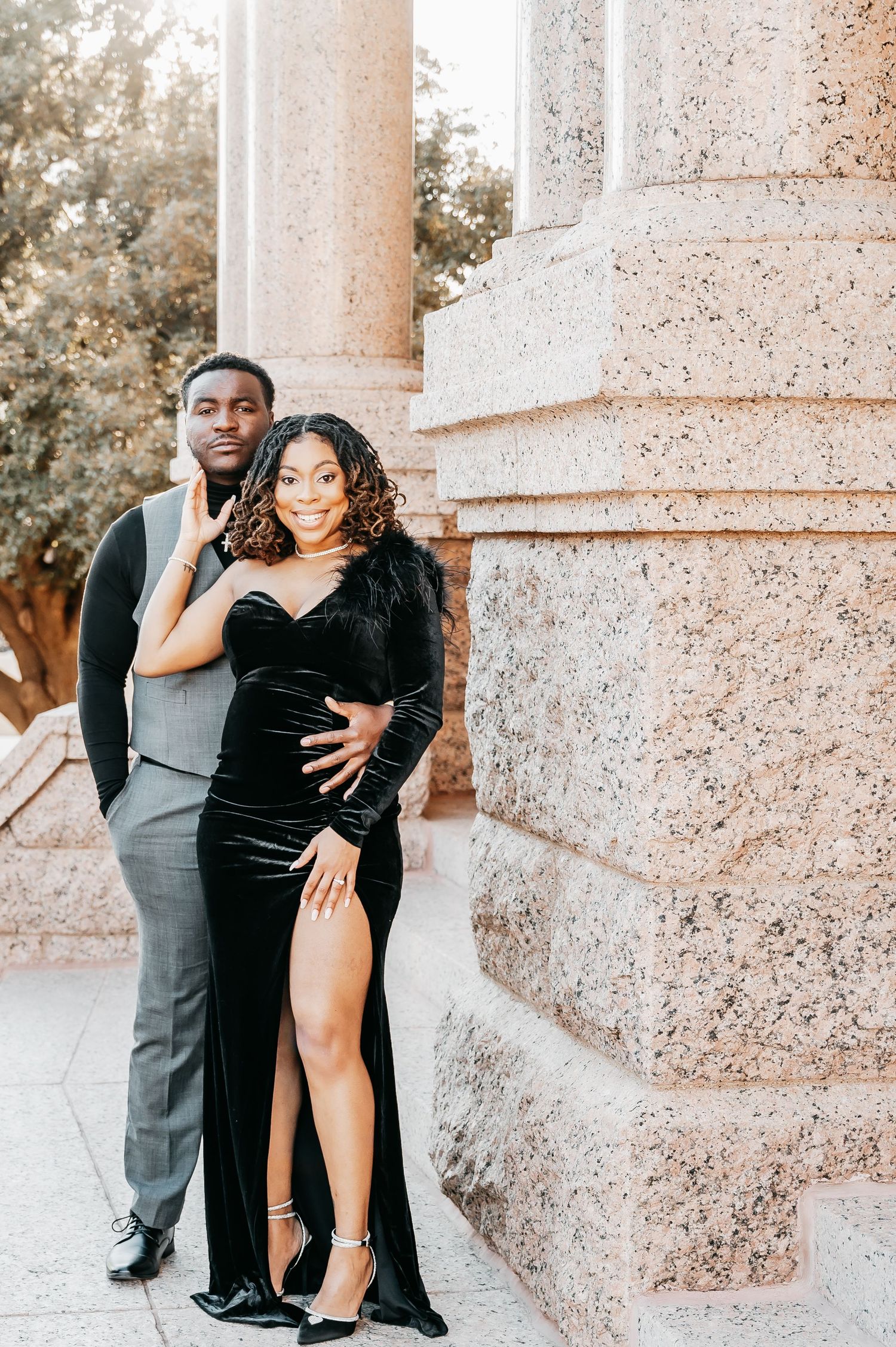 Elegant couple posing by stone columns, woman in black velvet gown with slit, man in gray suit, formal portrait.