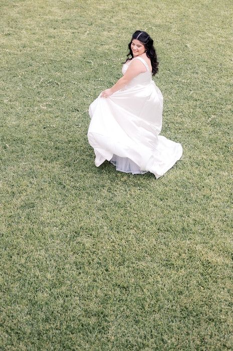 Bride twirling in white ball gown on green grass, lifting skirt while smiling outdoors.