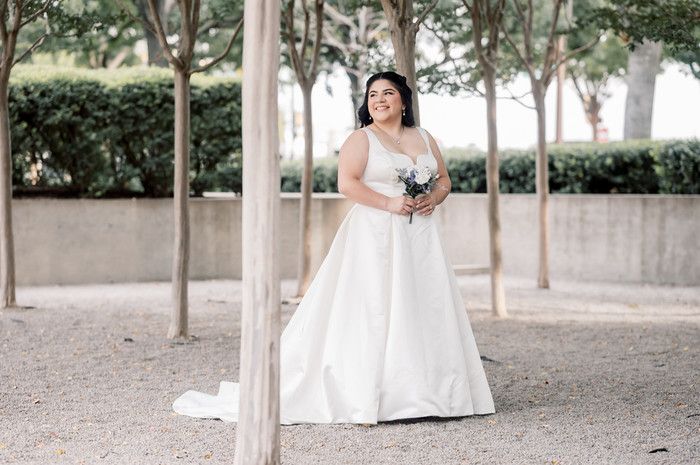 Bride in classic white A-line gown with train holds bouquet while standing between trees outdoors.