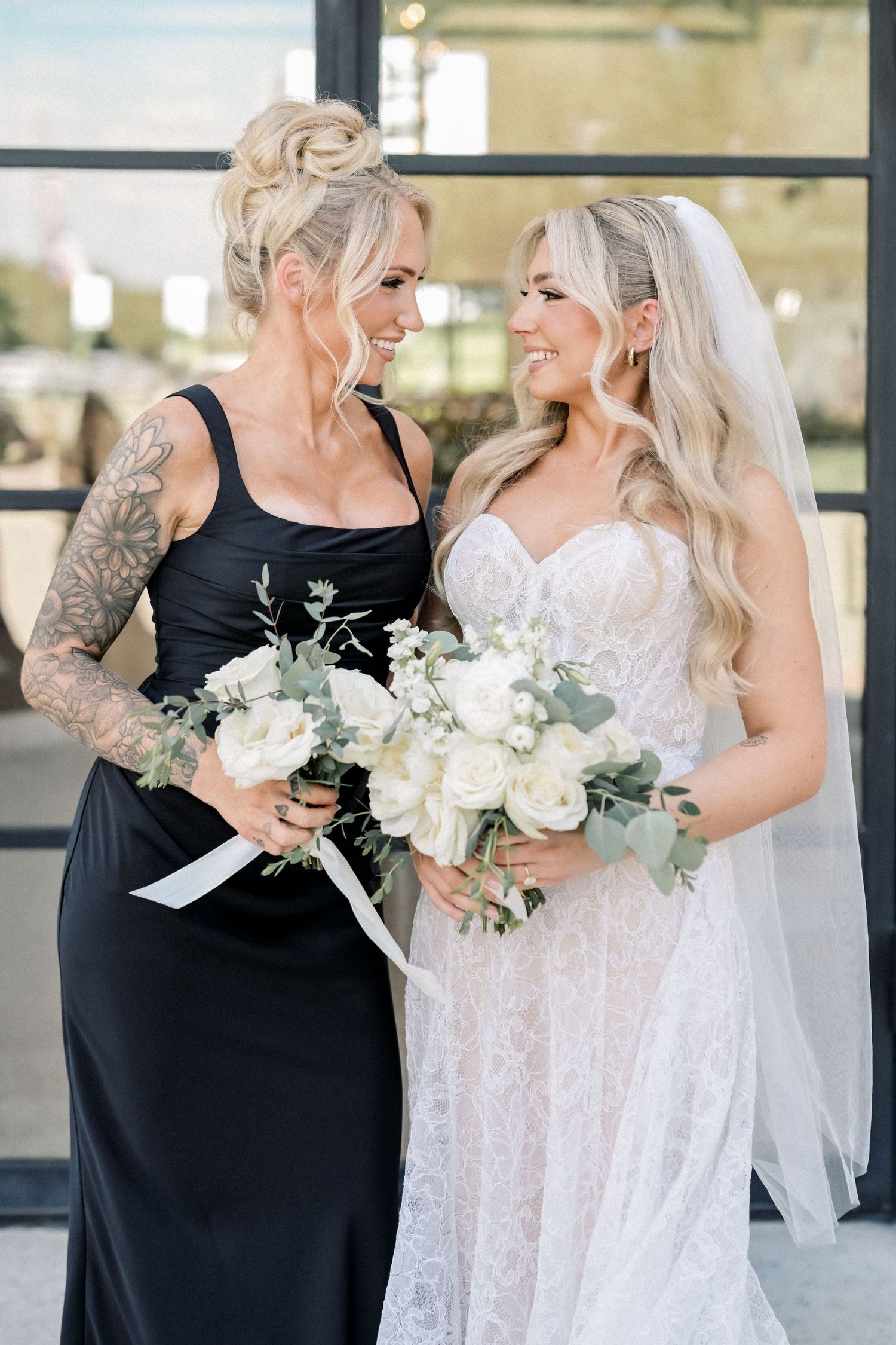 Bride in lace gown and bridesmaid in black dress holding white floral bouquets, smiling at each other outdoors.