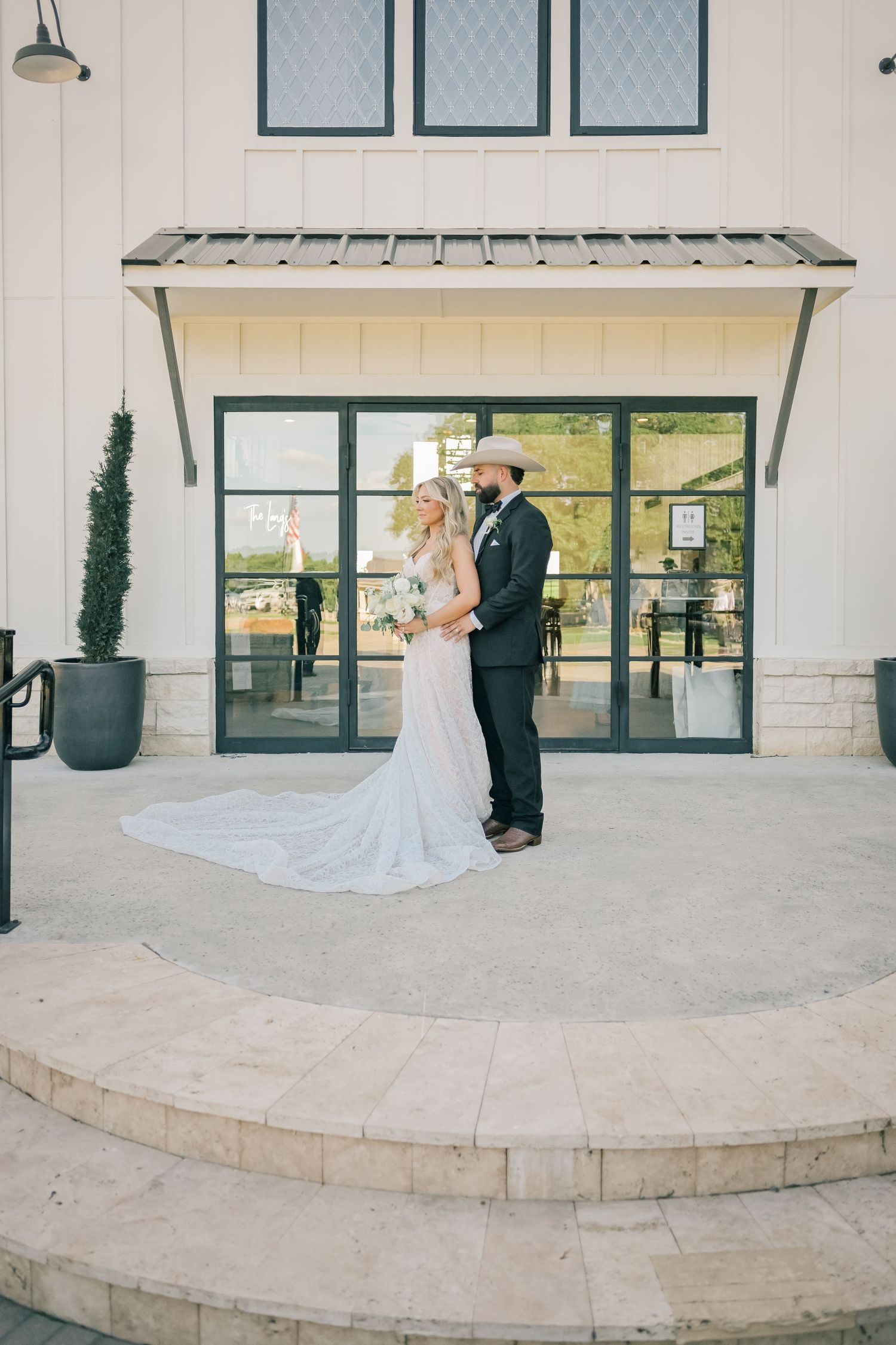 Bride and groom in cowboy hat share romantic moment outside white barn wedding venue entrance.