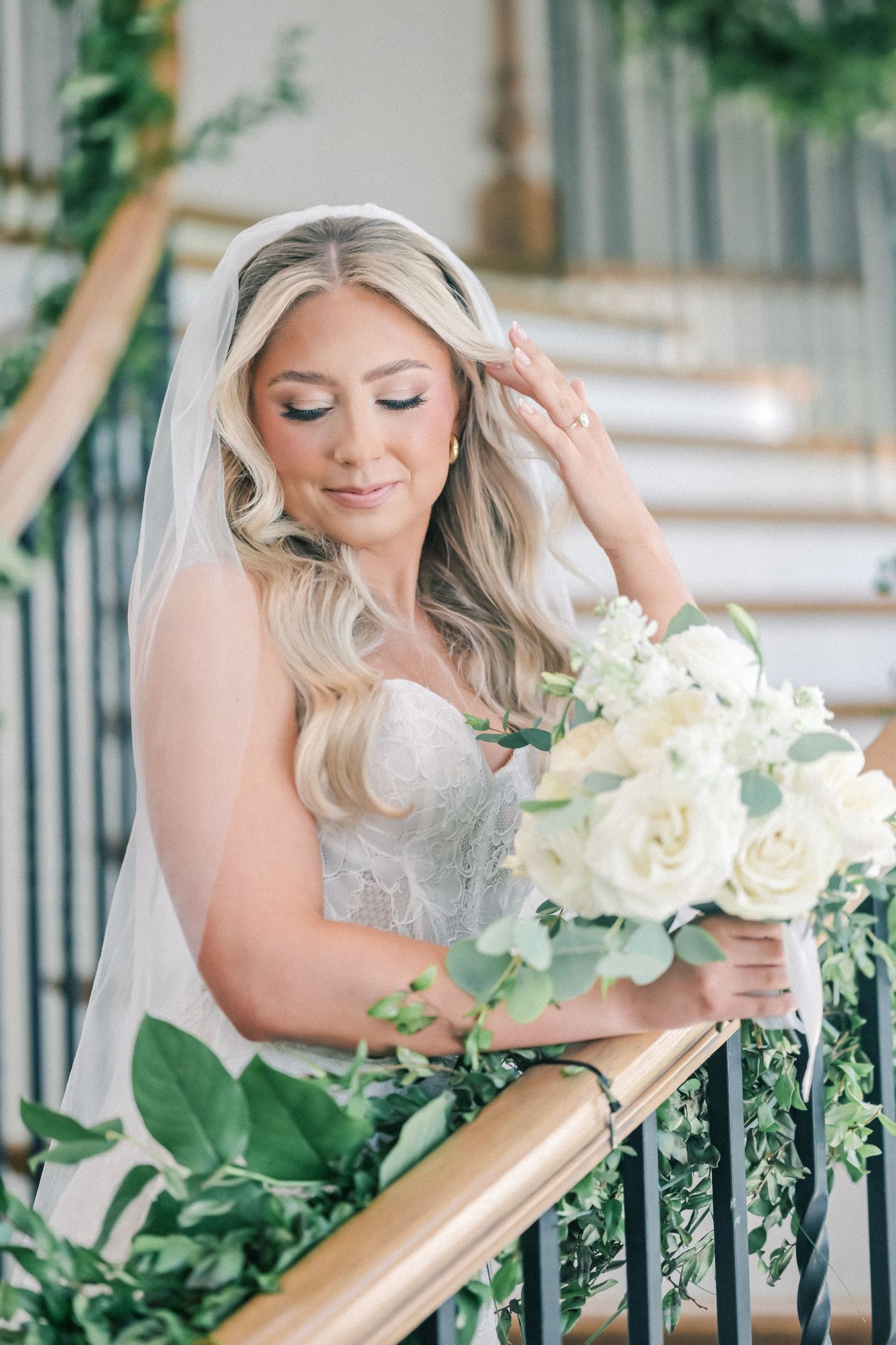 Elegant bride in lace gown holding white rose bouquet on decorated staircase.