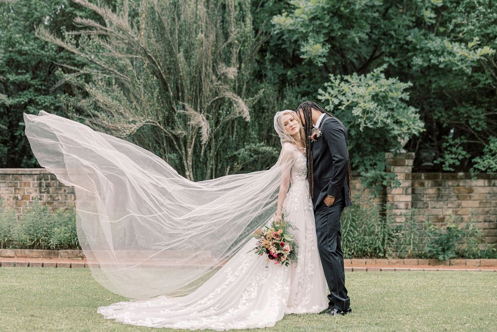Bride and groom share romantic kiss outdoors, her lace gown and veil flowing in the wind against lush garden backdrop.