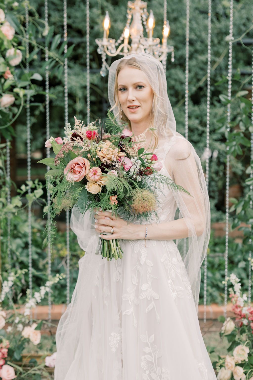 Blonde bride in lace gown holds colorful bouquet of pink roses and greenery before a chandelier backdrop.