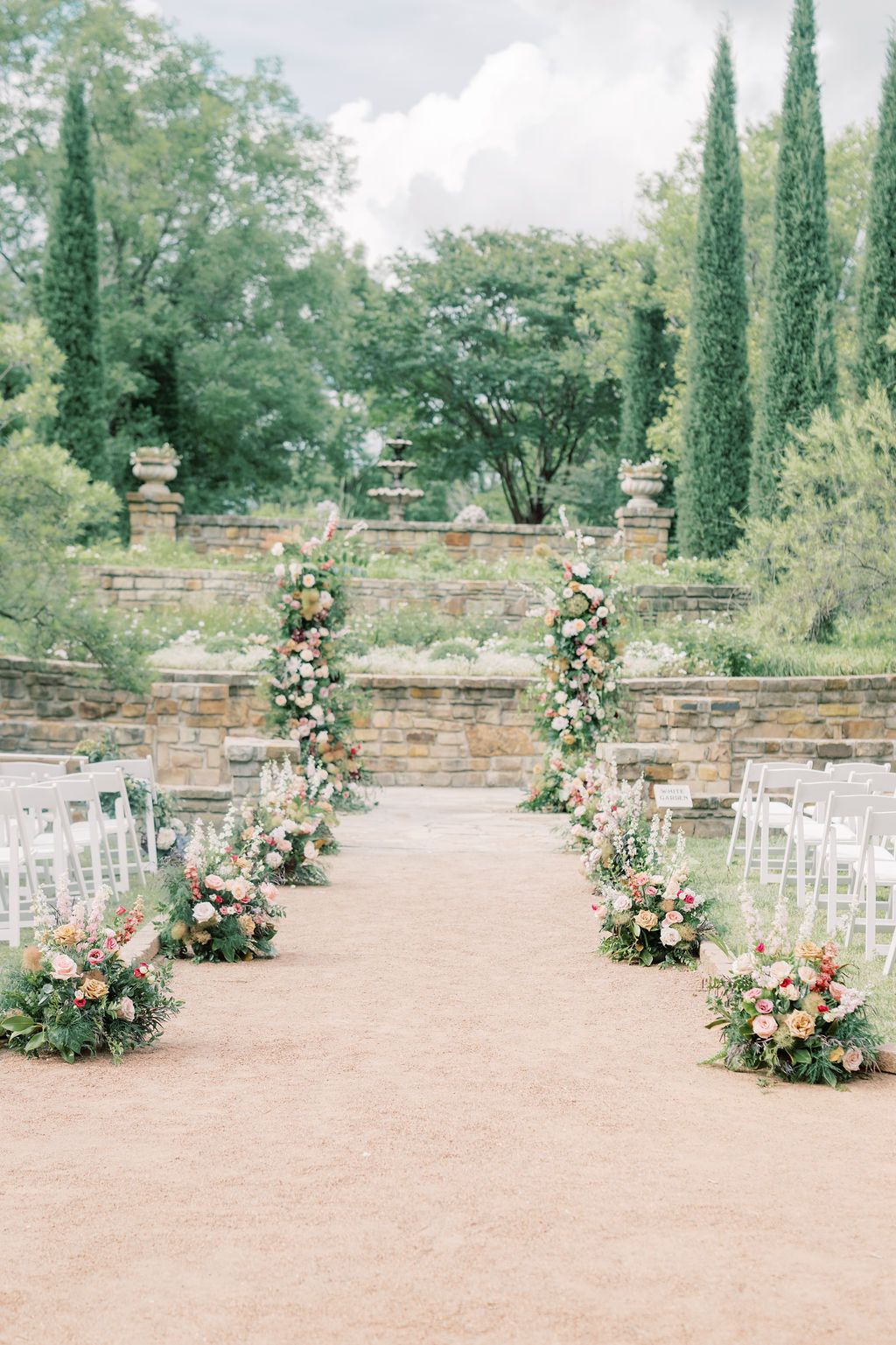 Elegant outdoor wedding ceremony aisle with floral arrangements, white chairs, stone walls, and cypress trees in a garden setting.