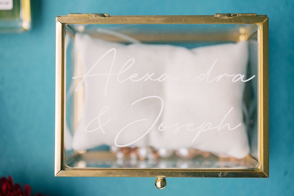 Gold glass ring box engraved with Alexandra & Joseph in white script on teal background.