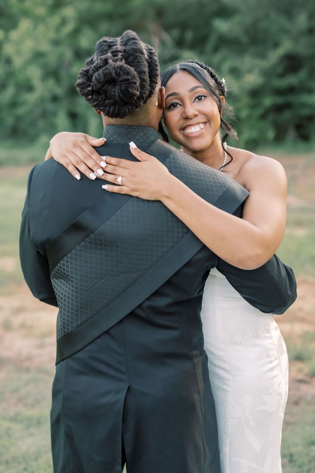 Bride in white gown smiles while embracing groom in black suit outdoors at wedding ceremony.