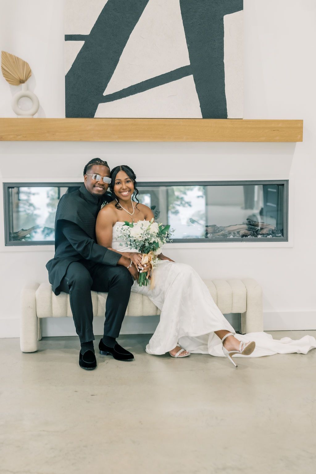 A bride and groom sit together smiling, she holds a white bouquet, wearing a lace gown, he wears a dark suit.