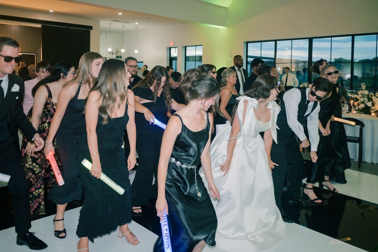Bride in white gown dancing energetically with guests in black dresses on a white dance floor at a wedding reception.