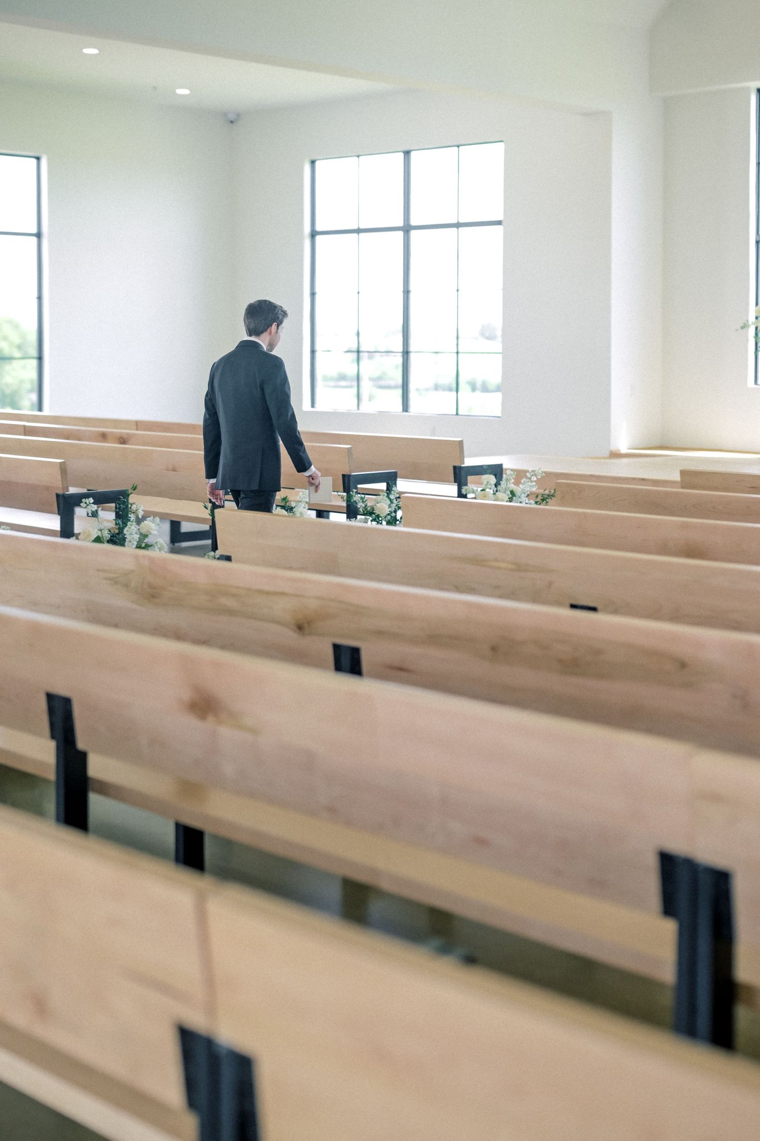 Groom in black suit walking alone between wooden pews in a bright modern white chapel before the wedding ceremony.