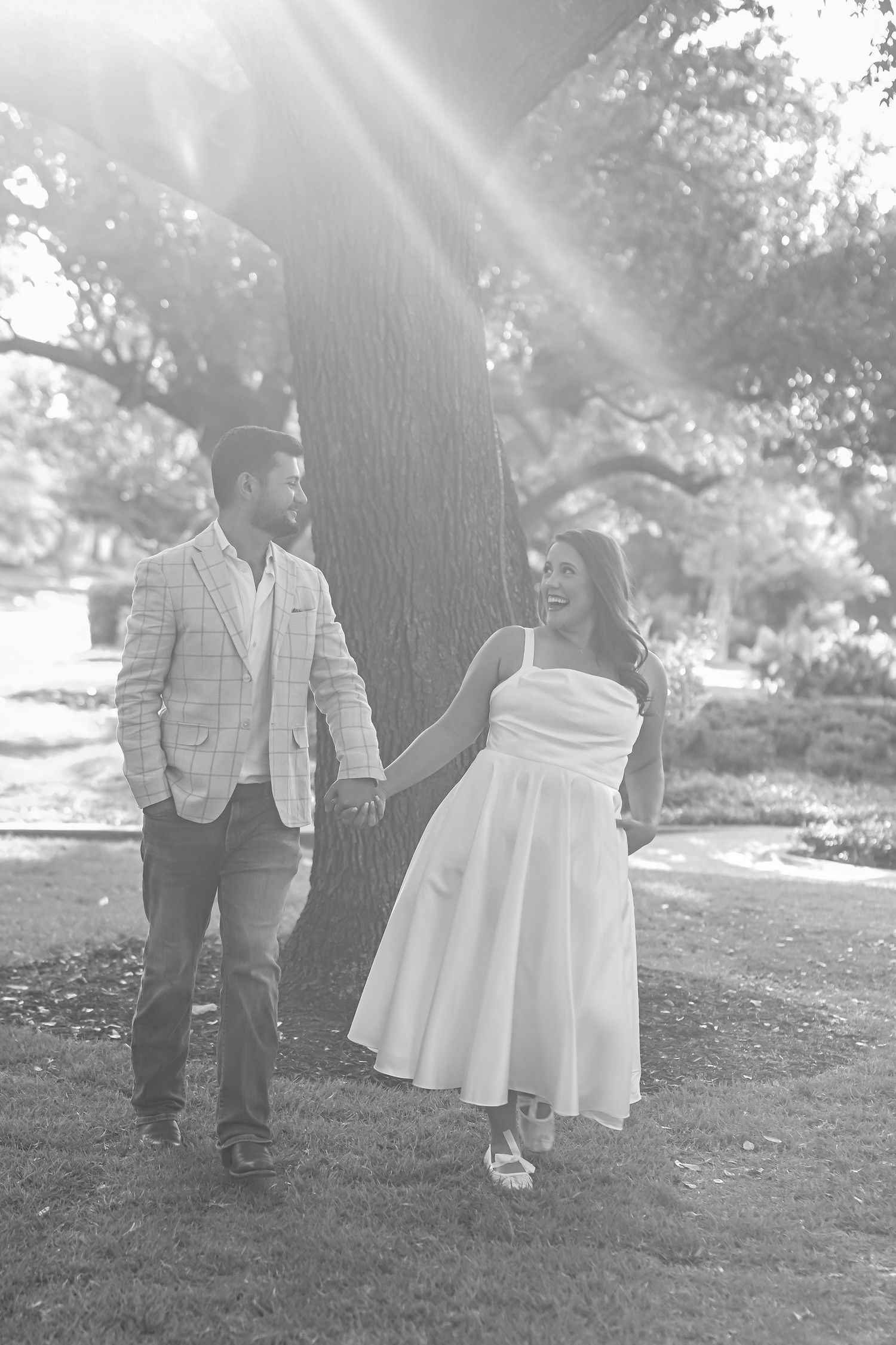 Black and white photo of a laughing couple holding hands and walking beneath a large oak tree with sunbeams streaming through.