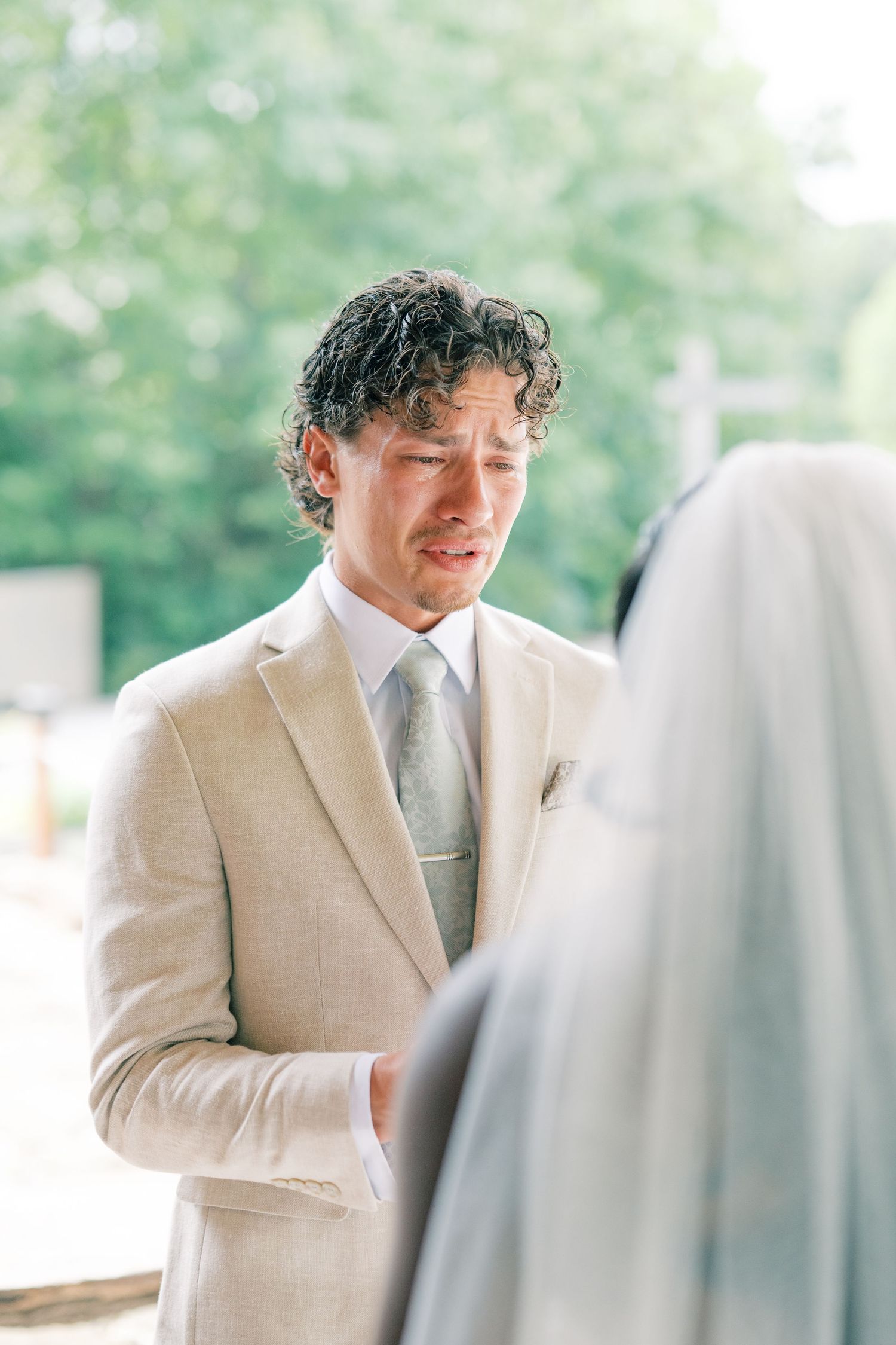 Emotional groom in tan suit with gray tie listens to wedding vows during outdoor ceremony.