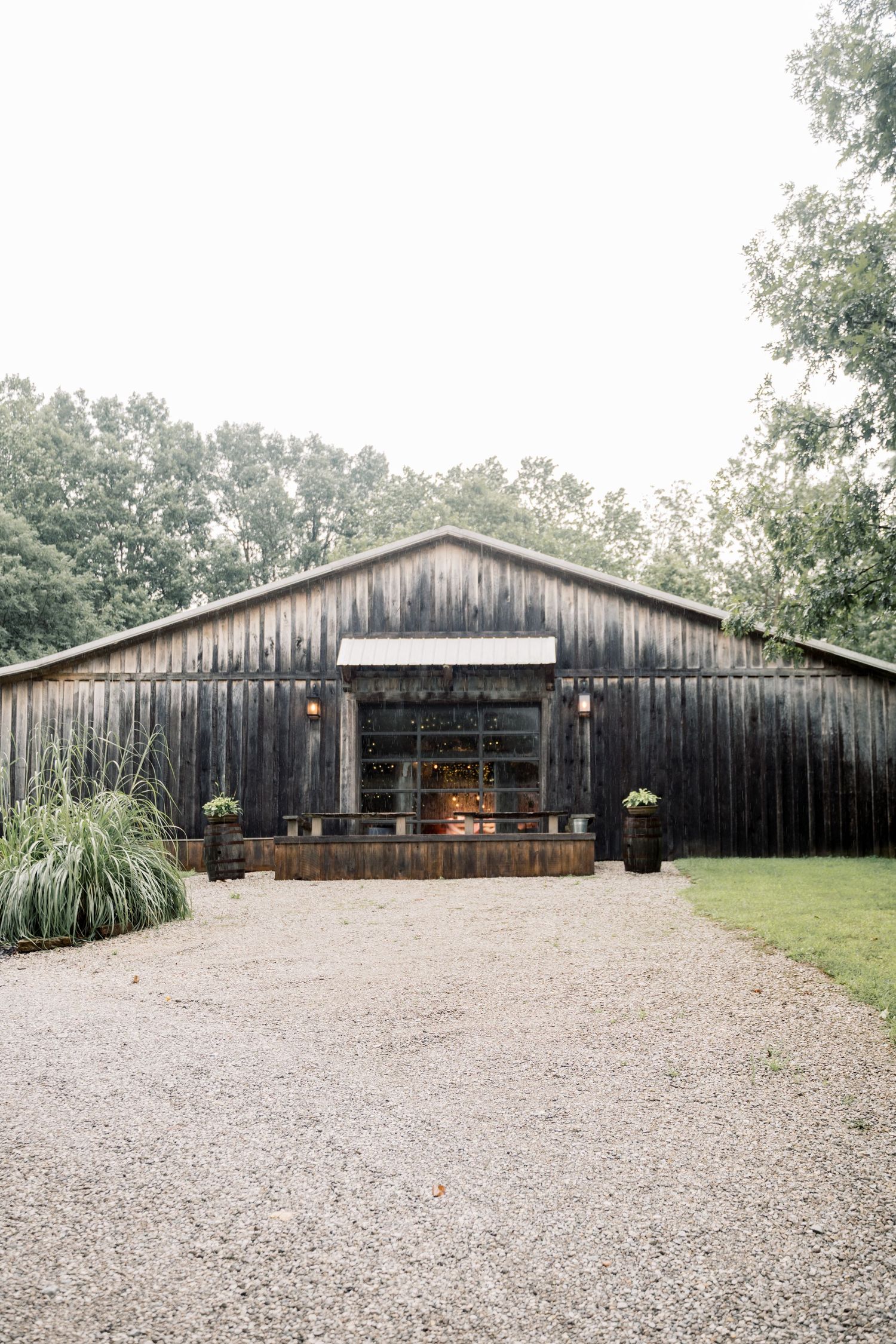 Rustic dark wood barn wedding venue with gravel driveway surrounded by lush green trees.