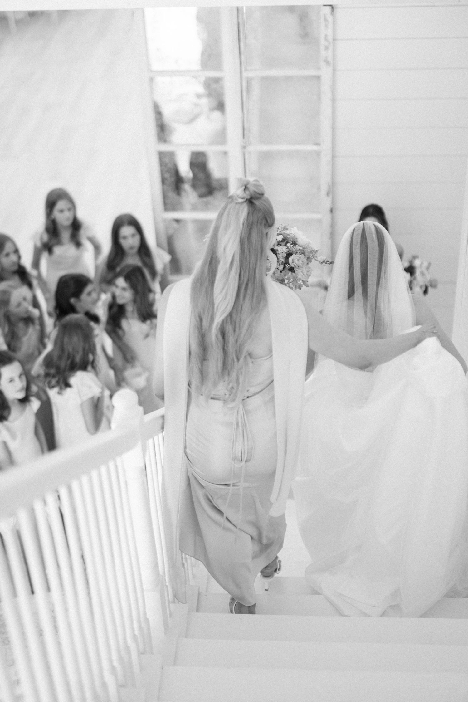 Black and white photo of bride descending staircase with bridesmaid as wedding guests gather below in white venue.