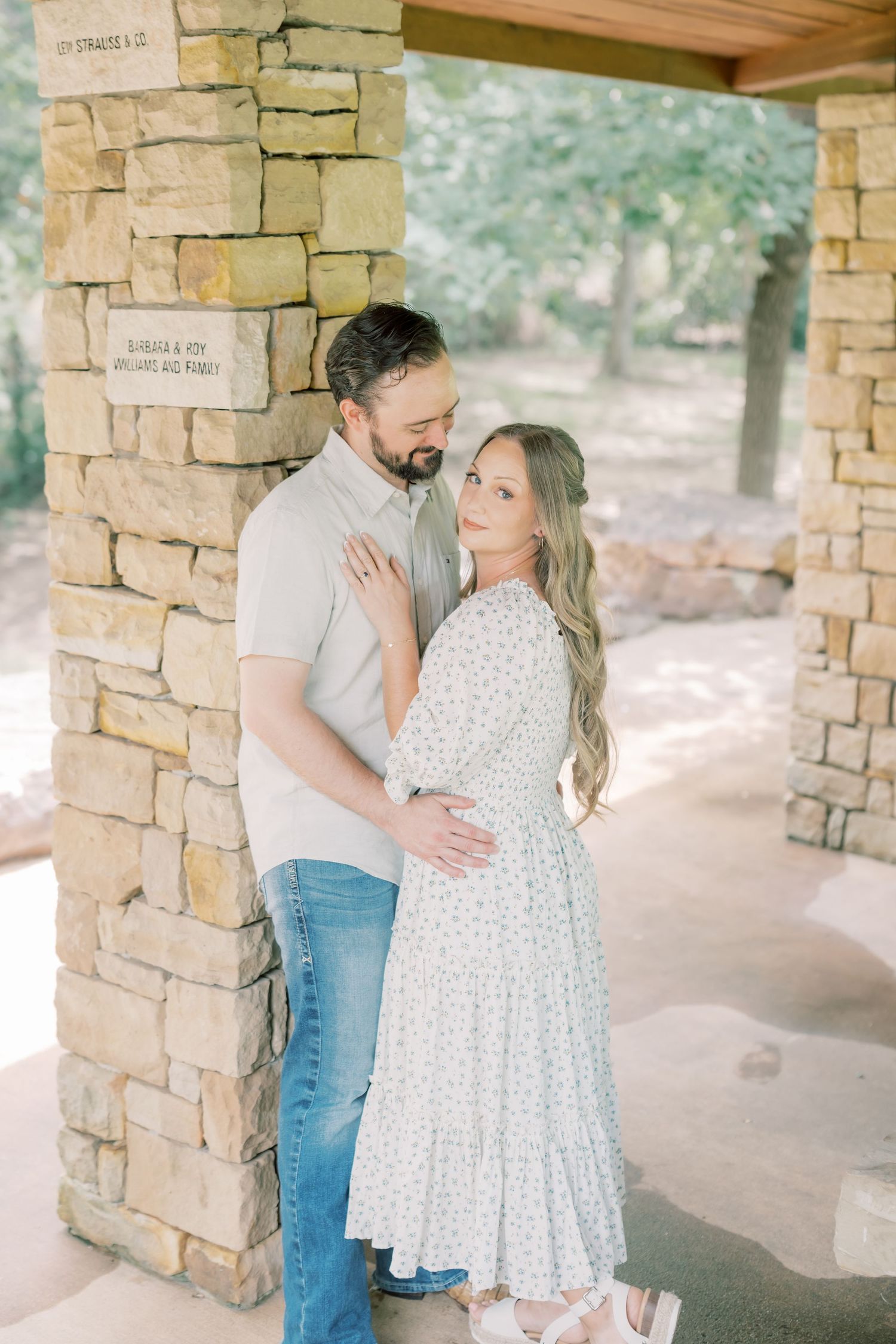Couple embracing near a rustic stone pillar in a shaded outdoor pavilion setting.