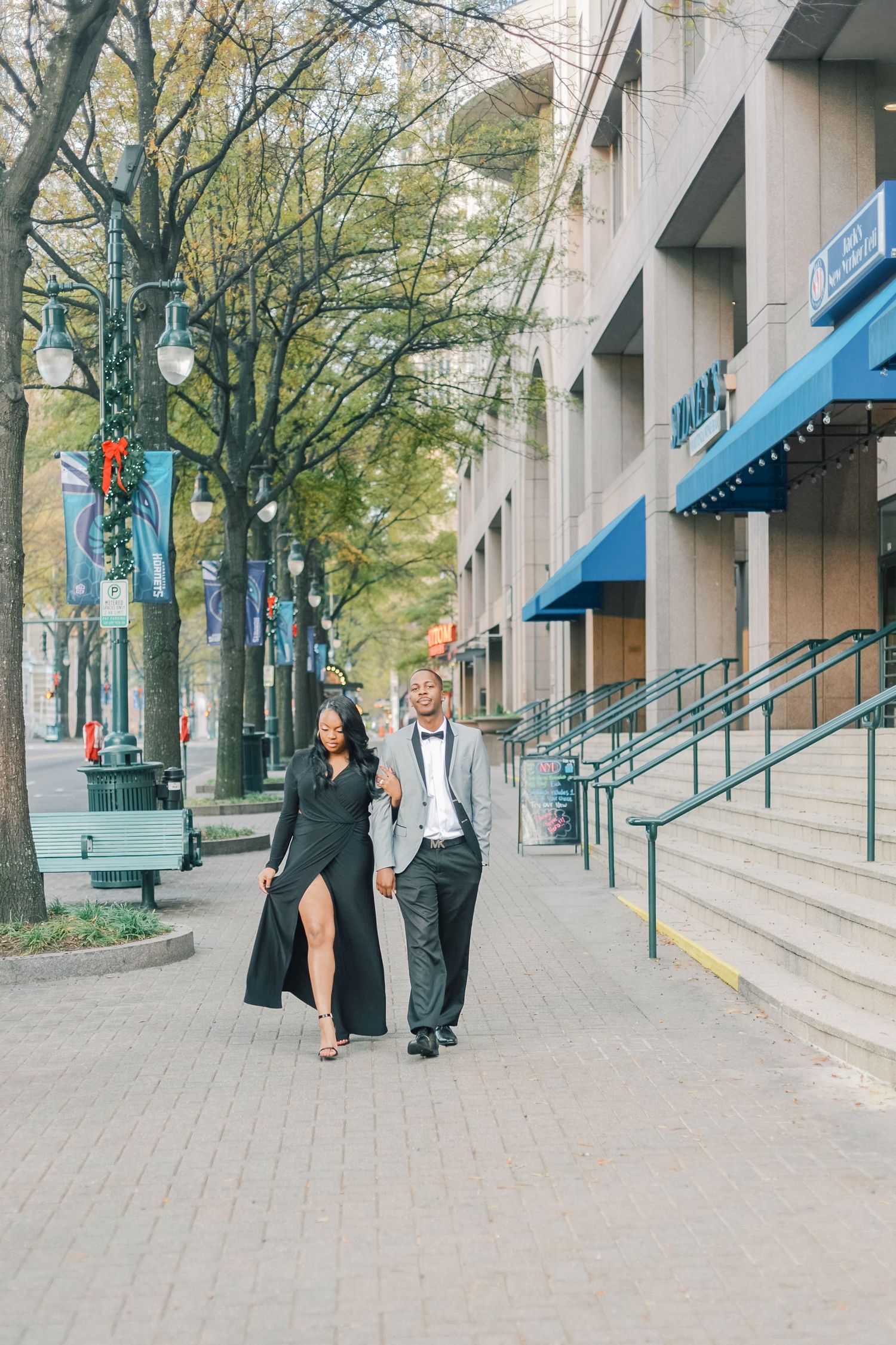 Elegant couple in formal wear walking hand in hand along a city sidewalk with fall trees.