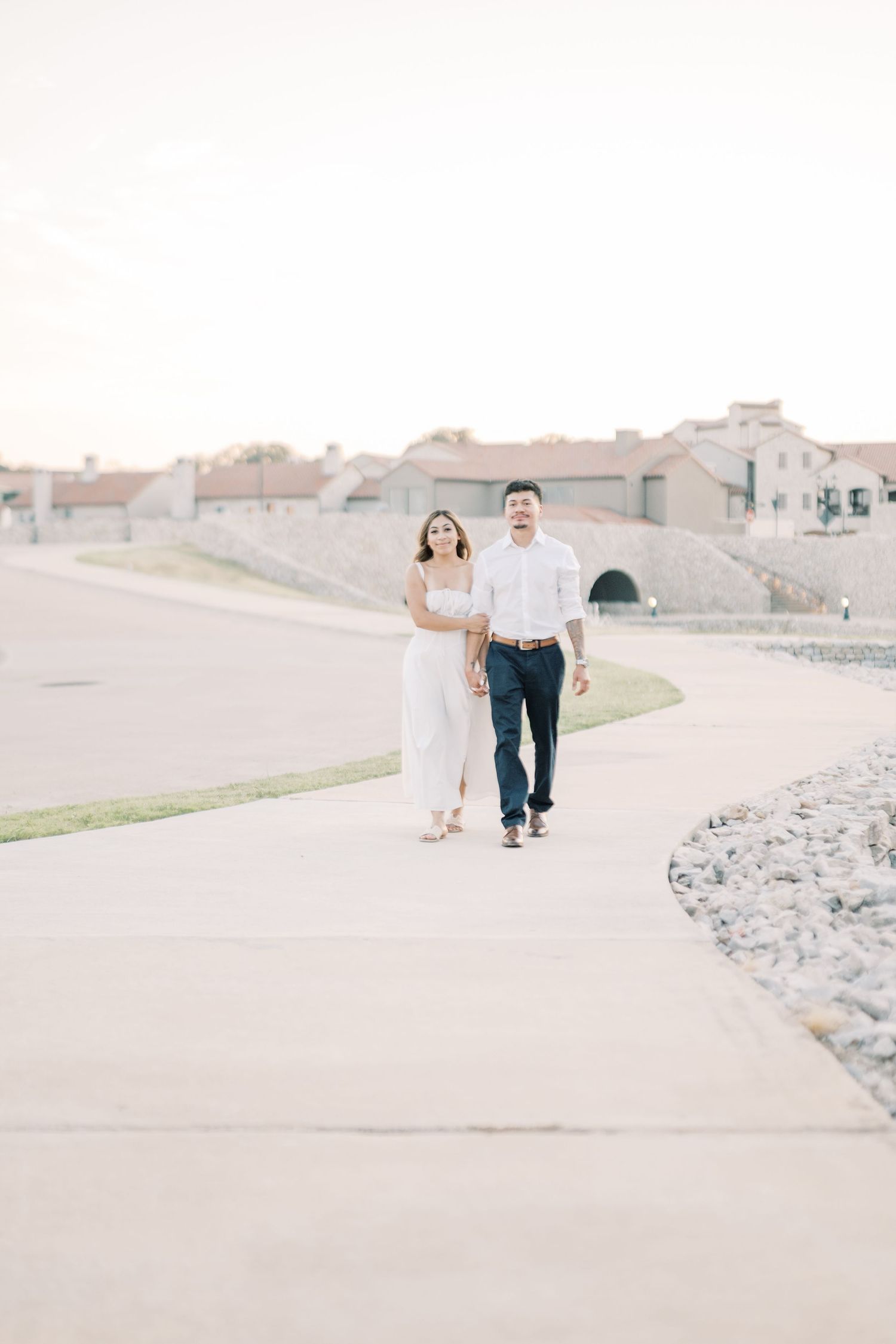 Couple in white outfits walking hand in hand along scenic outdoor pathway.