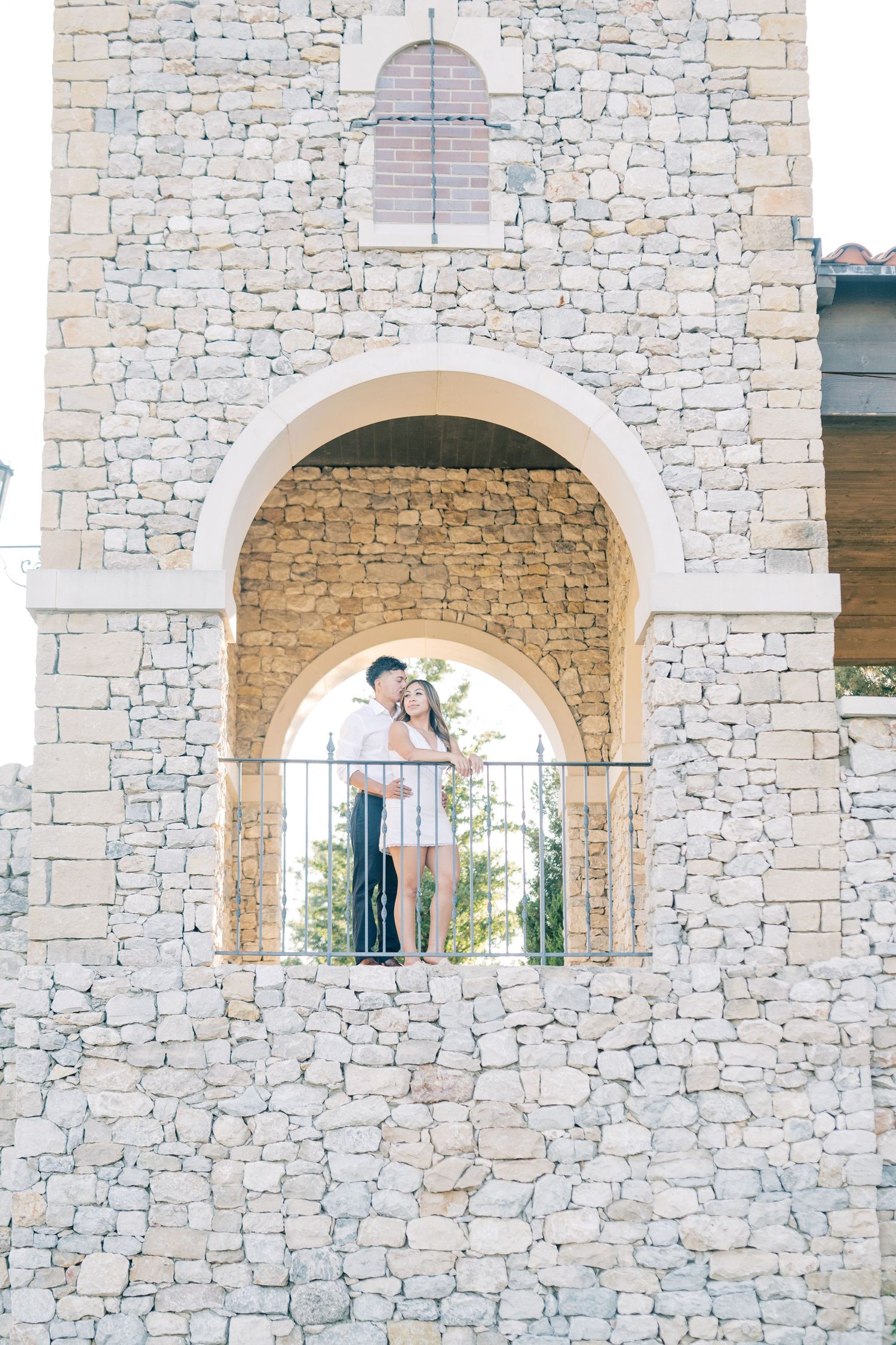 A romantic couple shares a kiss under a beautiful stone arch during their outdoor engagement session.