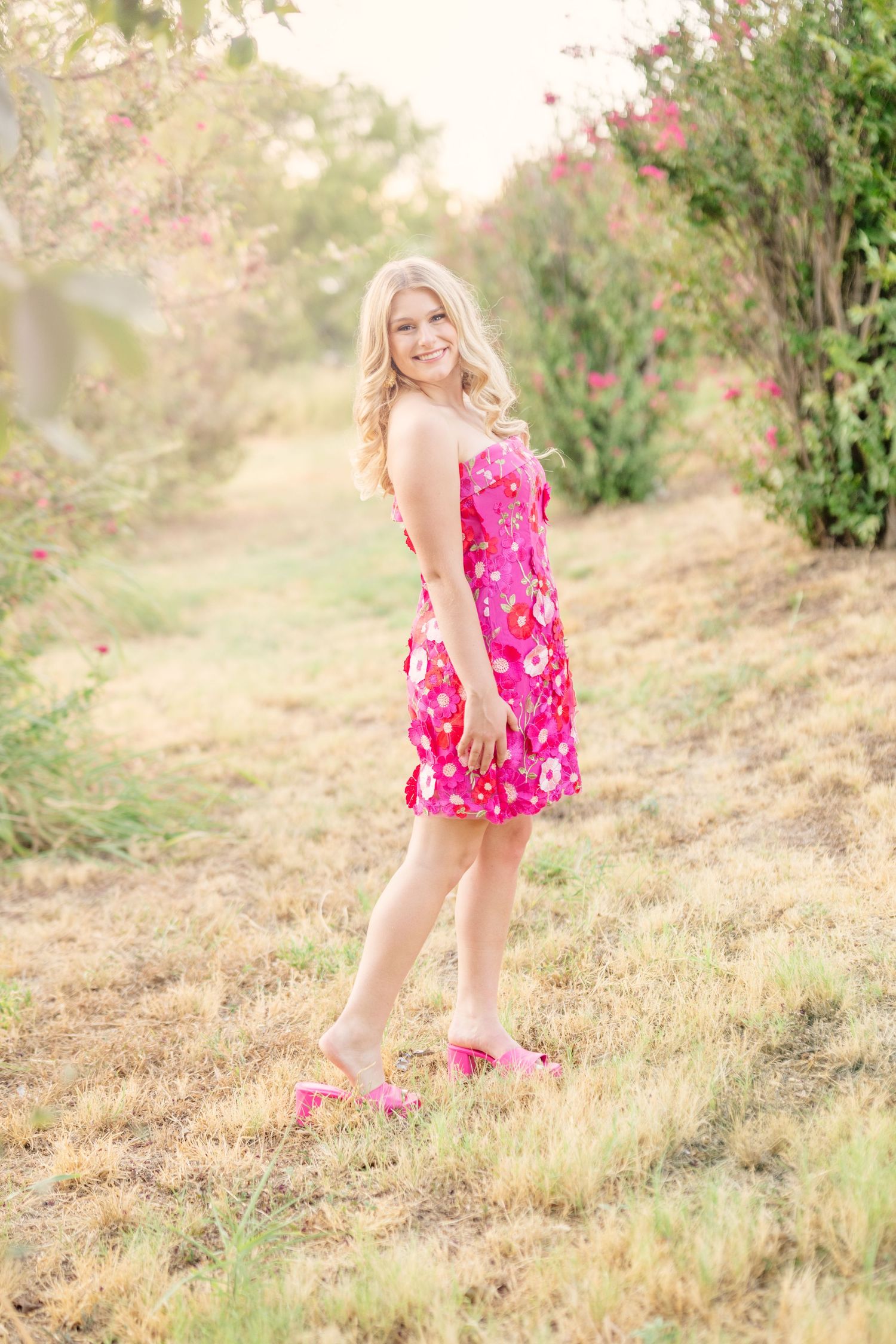 Young blonde woman in pink floral sundress and heels smiling outdoors in a garden path with blooming flowers.