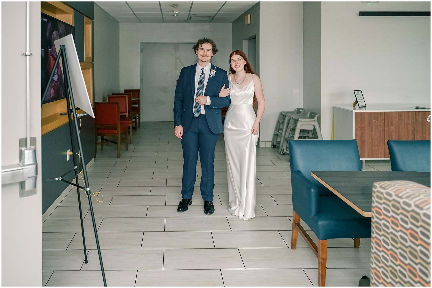 A wedding couple poses in a modern office space with white walls and tiled floors.