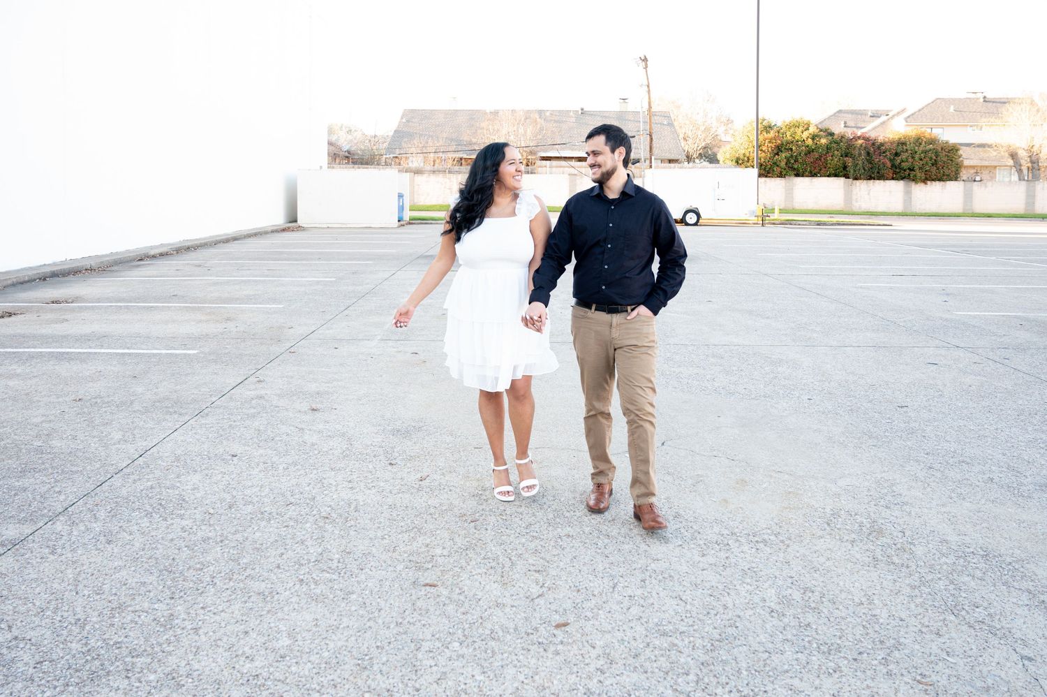 A couple walking together on a concrete surface, with the woman in a white dress and the man in a navy shirt and khakis.