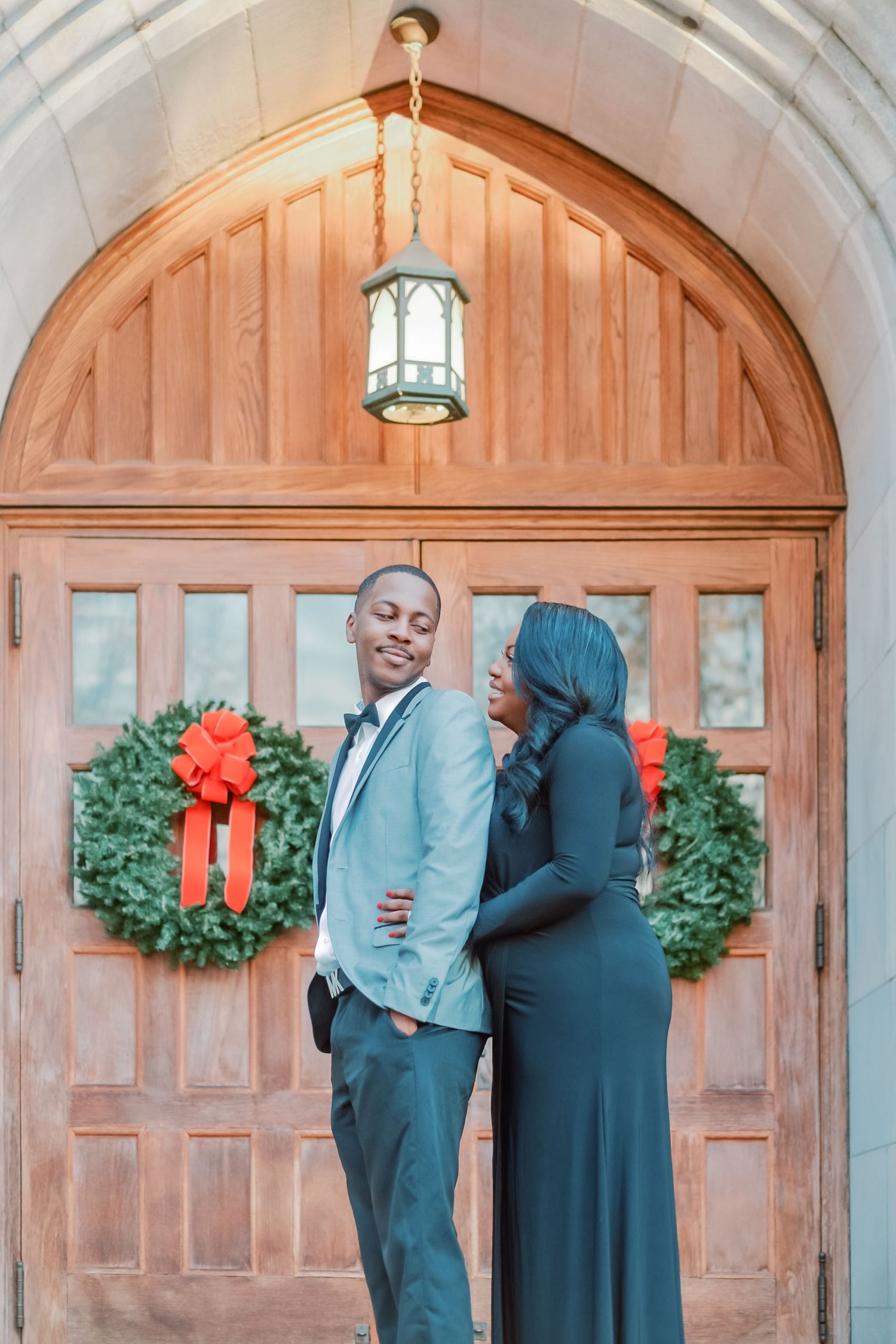 Couple posing in front of wooden church doors with Christmas wreaths, man in gray suit, woman in black gown.