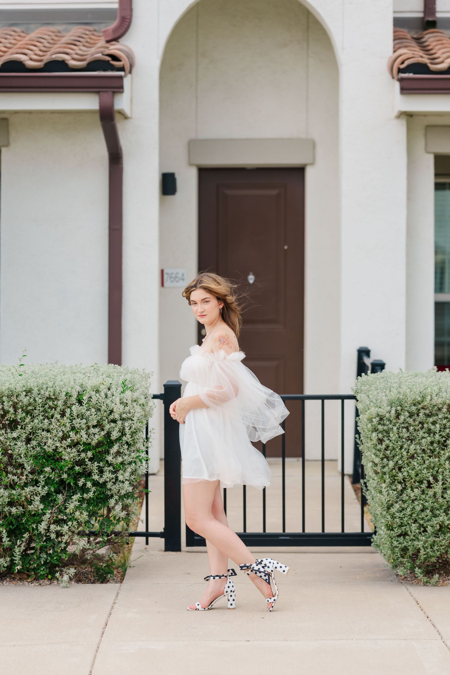 Young woman in white off-shoulder dress and polka dot heels poses gracefully on sidewalk outside elegant building.