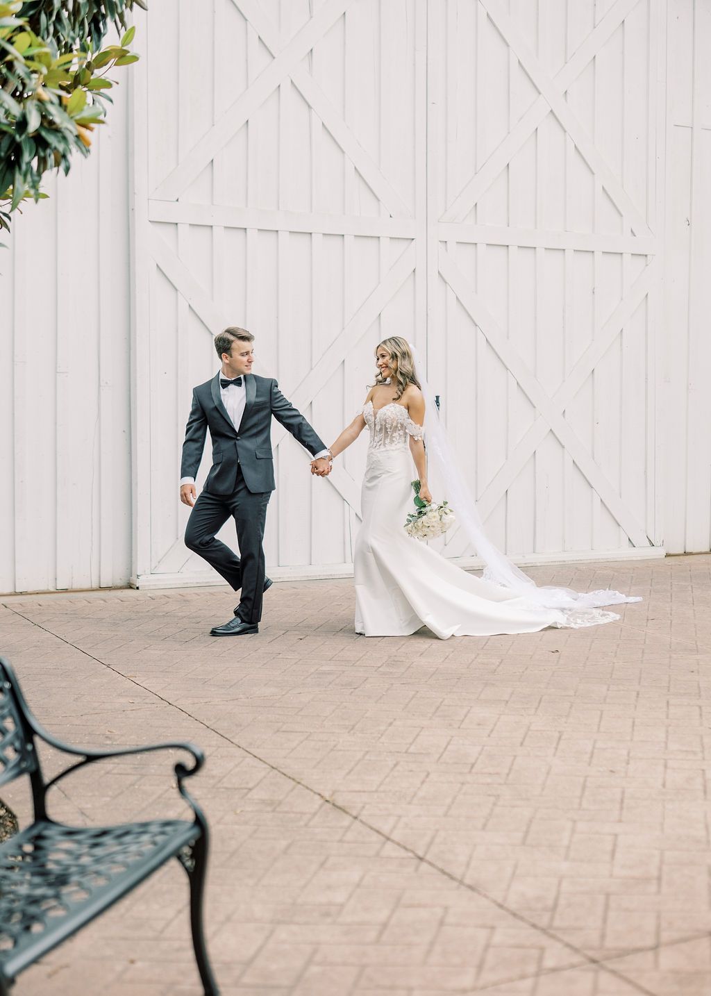 Bride and groom walk hand in hand before a white barn door, her flowing gown trailing behind on brick pavers.