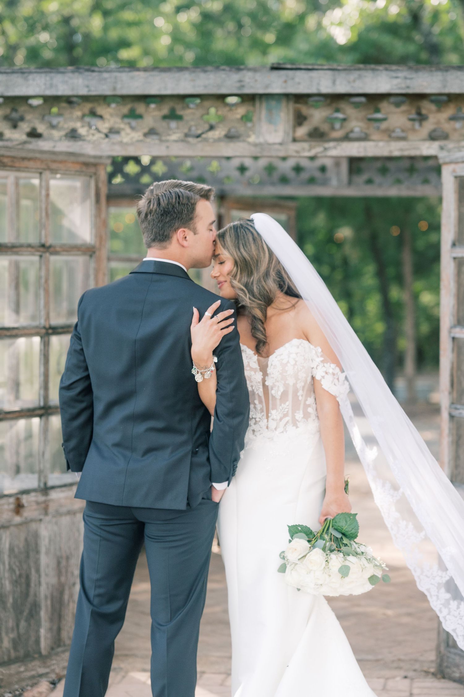 Bride and groom share a tender kiss outdoors near a rustic wooden arbor, bride holding white bouquet in elegant lace gown.