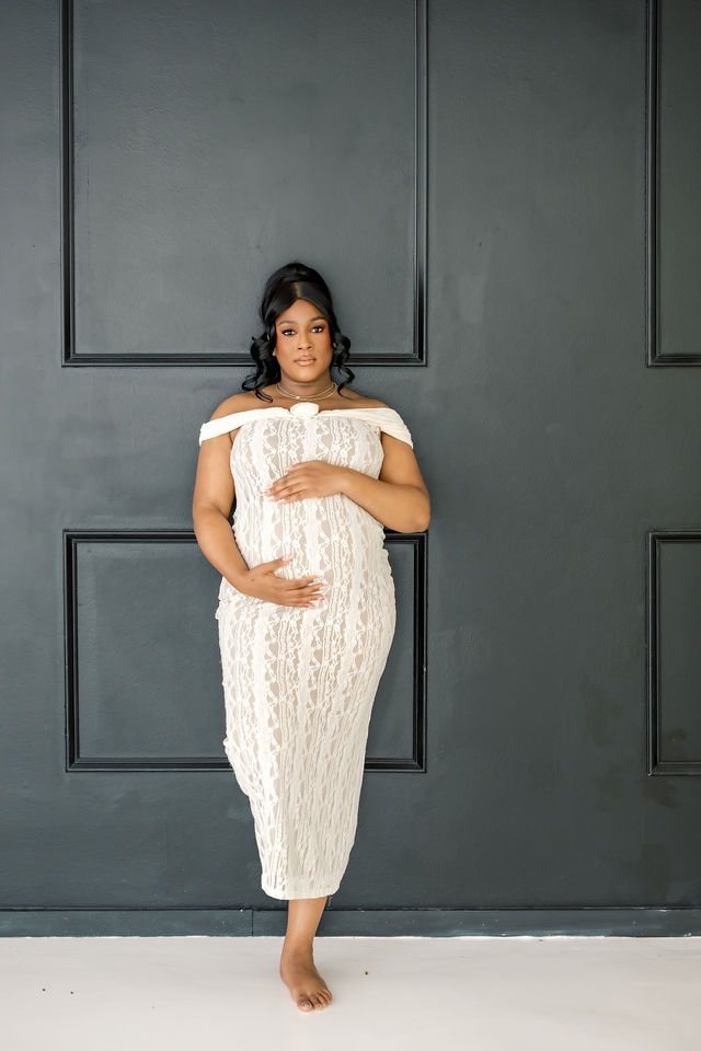 Pregnant woman in white lace off-shoulder dress posing against dark gray wall.