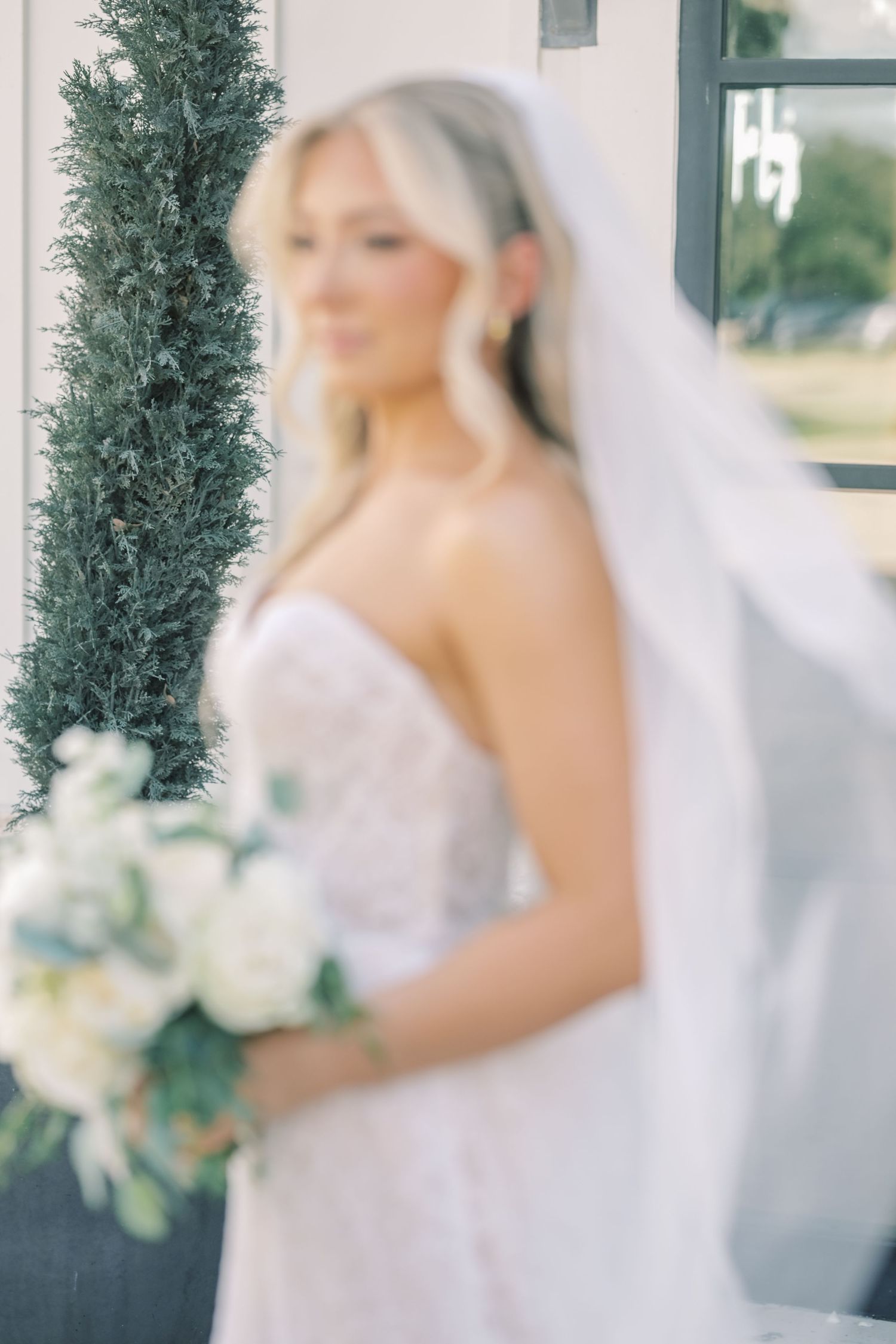 Soft focus portrait of blonde bride in strapless lace gown holding white rose bouquet with veil flowing.