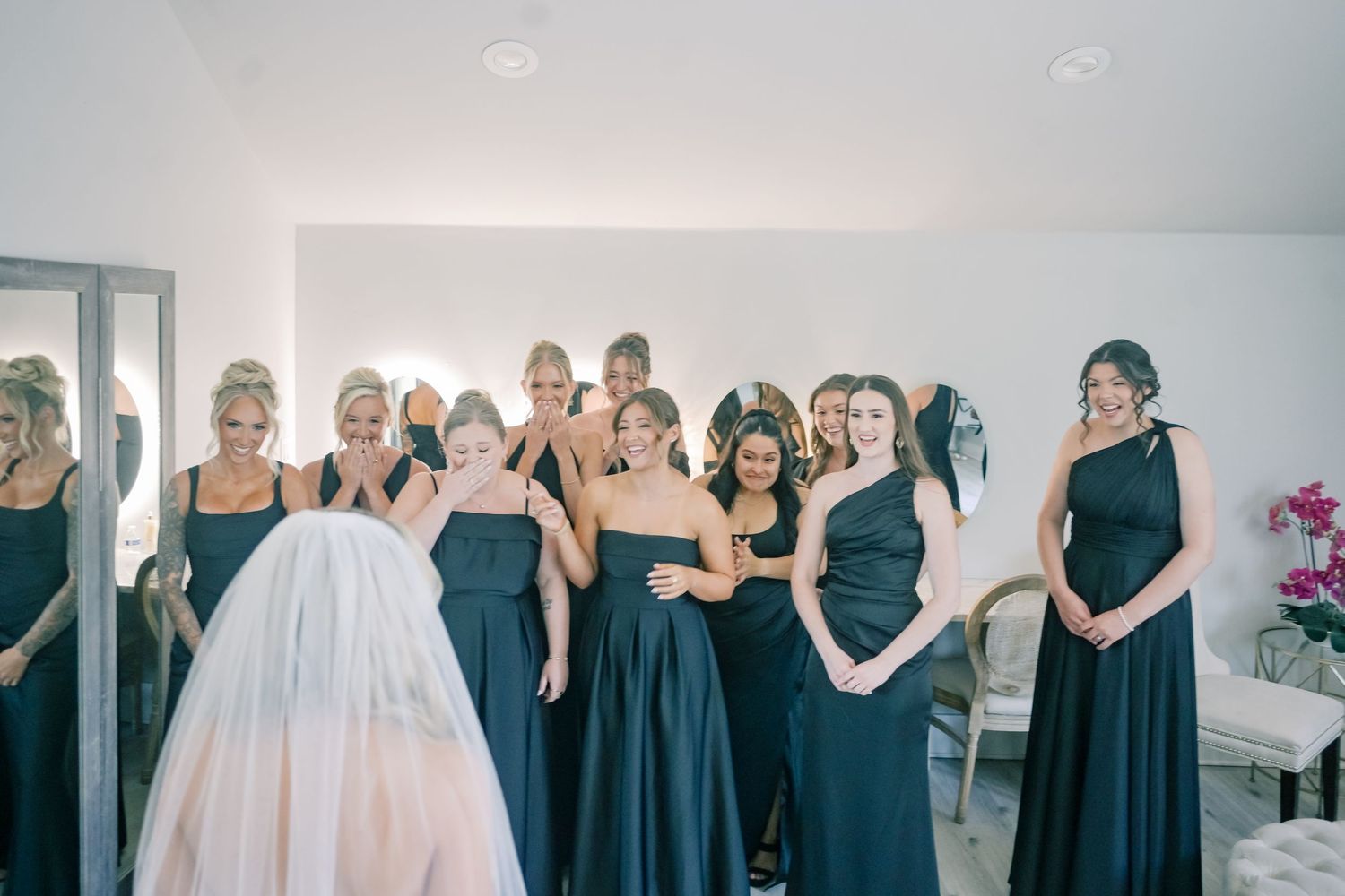 Bridesmaids in navy gowns react with joy as bride reveals wedding dress during getting ready moment.