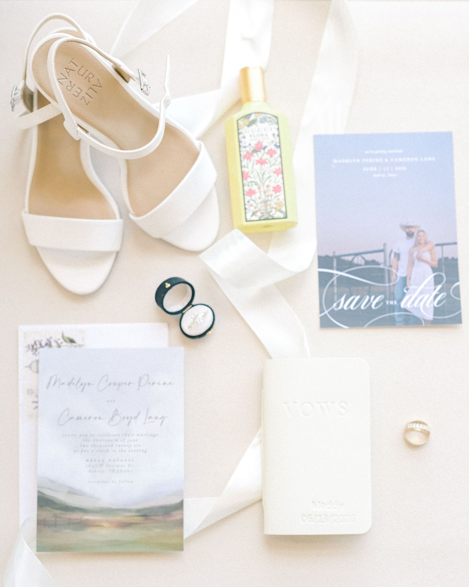 Elegant bridal flat lay featuring white heels, wedding invitation, save the date card, rings, and floral perfume bottle.