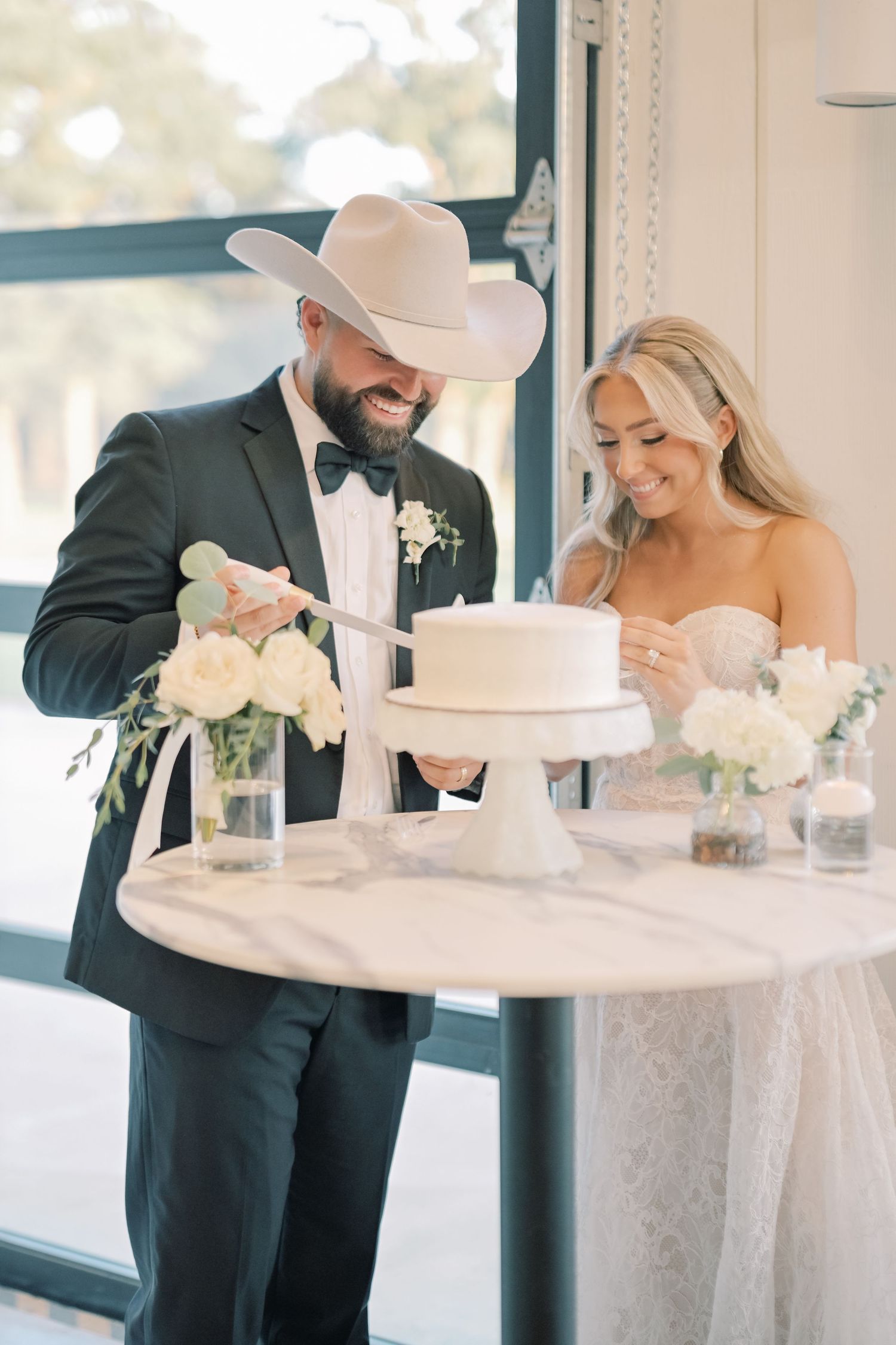 Laughing couple in cowboy hat cuts elegant white two-tier wedding cake at marble table.