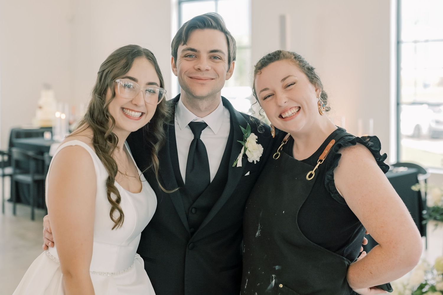 Bride in white dress and groom in black suit smiling with a guest wearing black ruffled top at wedding reception.