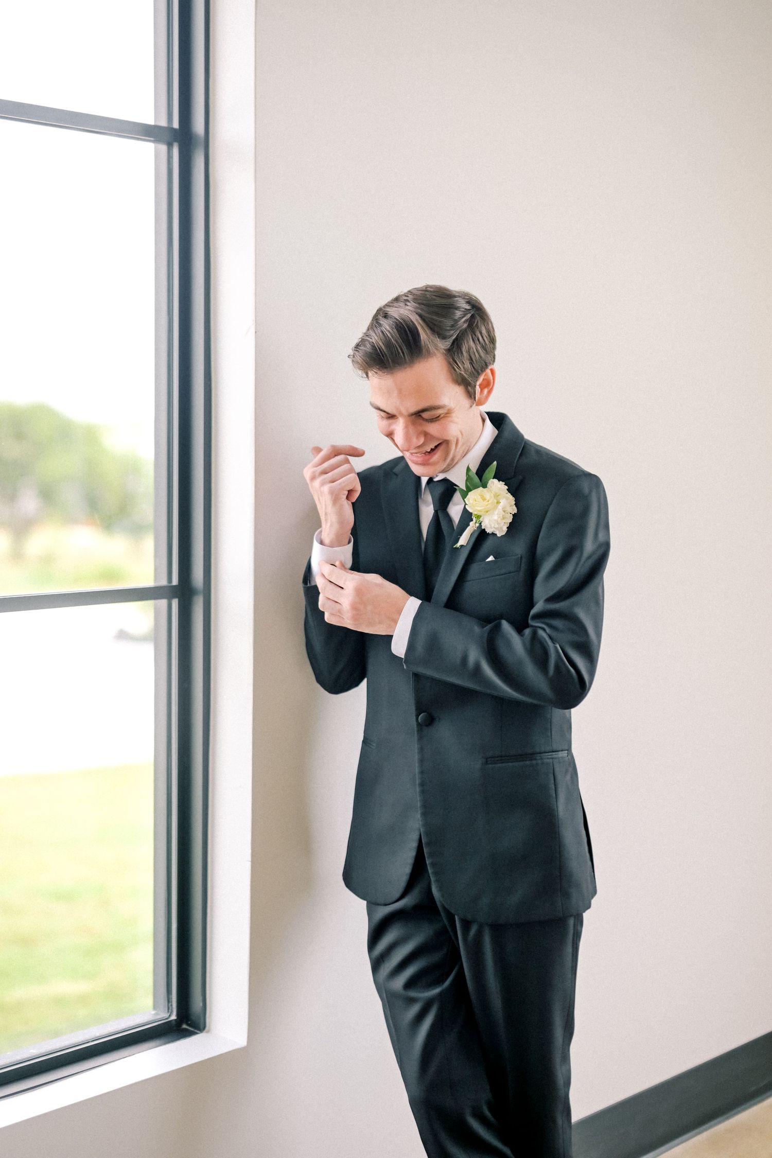 Dapper groom in dark suit adjusting cufflinks by window with boutonniere on lapel.