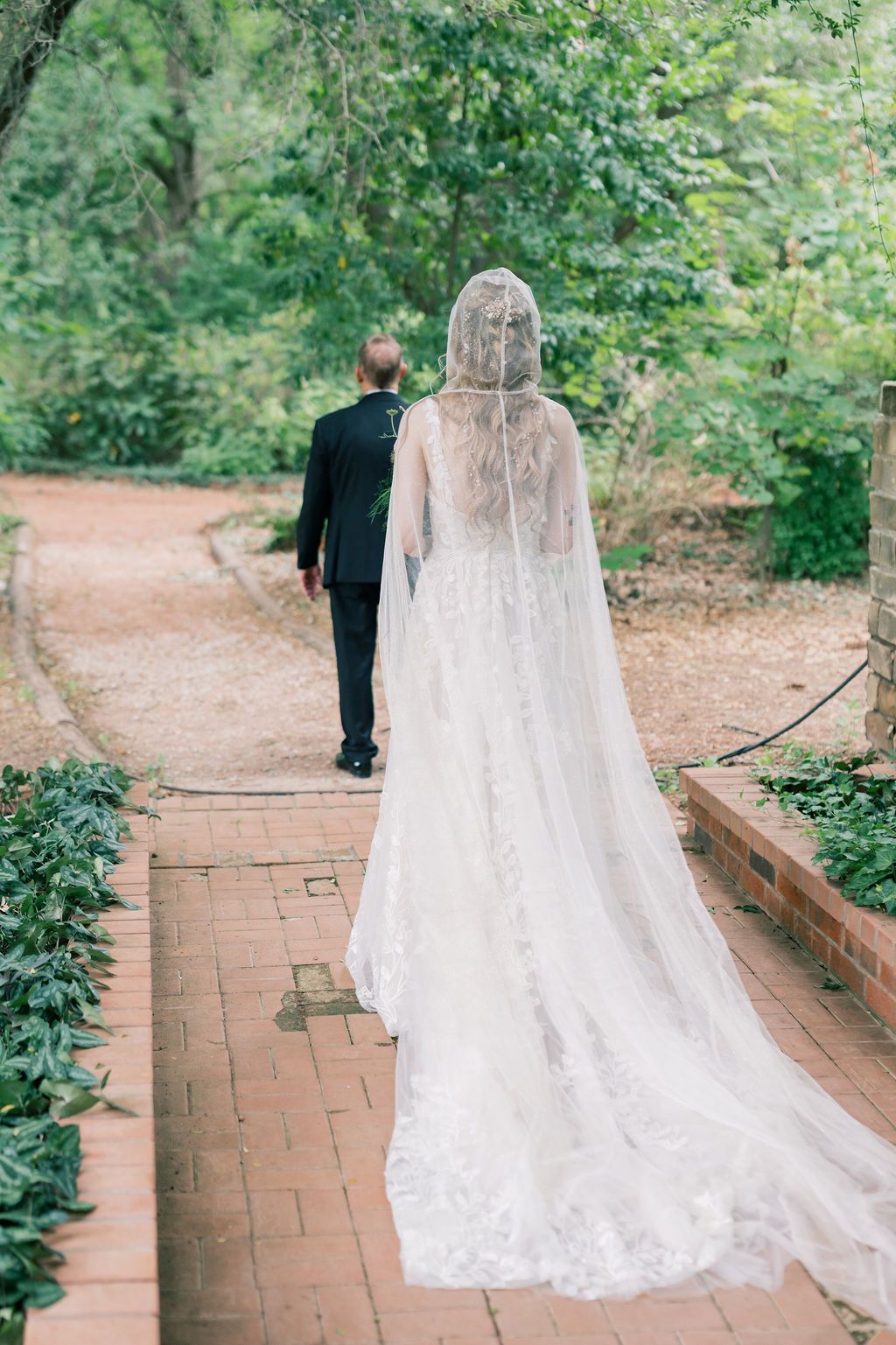 Bride in lace gown with cathedral veil walks with groom in black tuxedo along brick garden path.