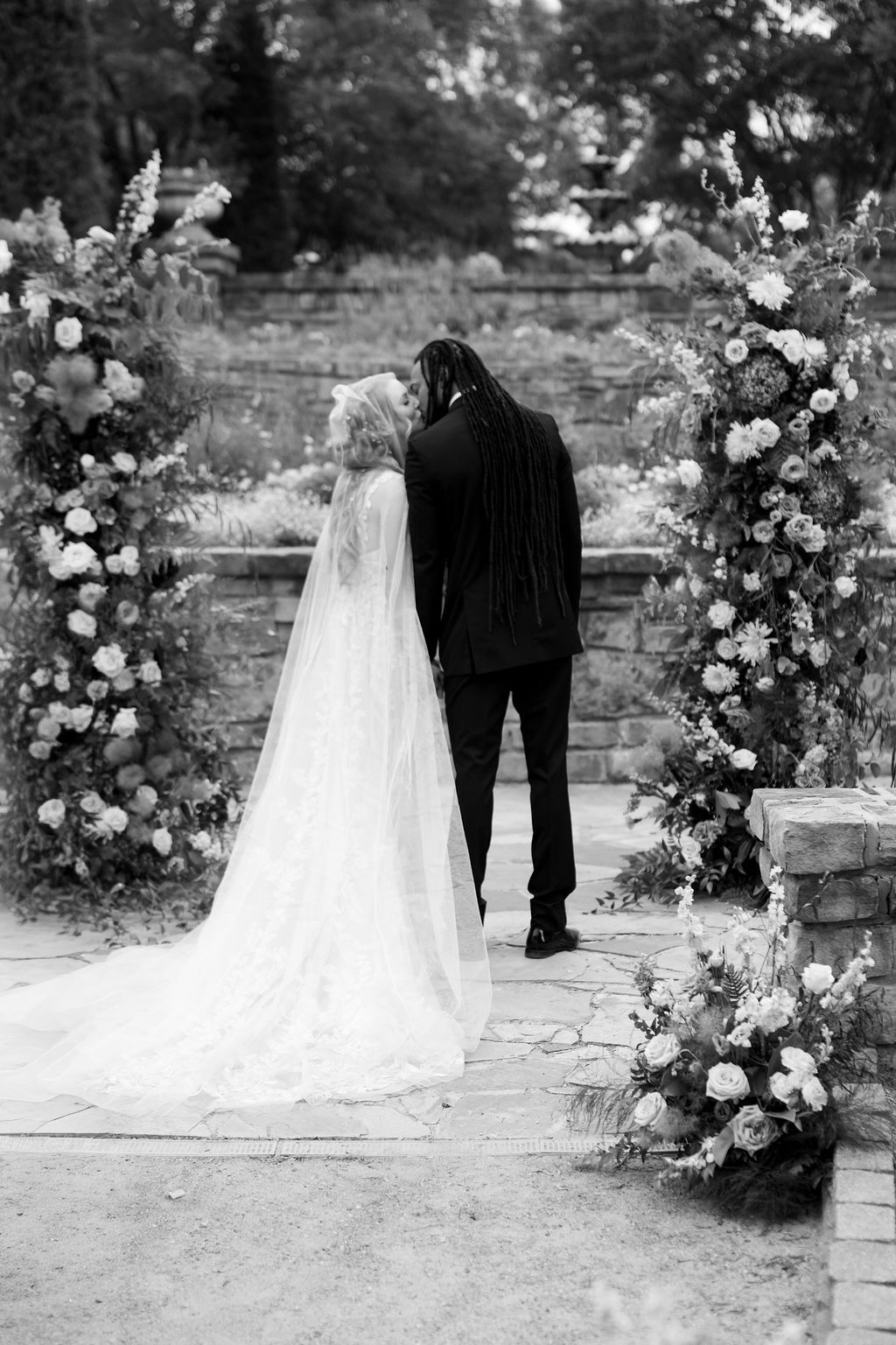Black and white photo of bride and groom standing at floral arch during outdoor wedding ceremony.
