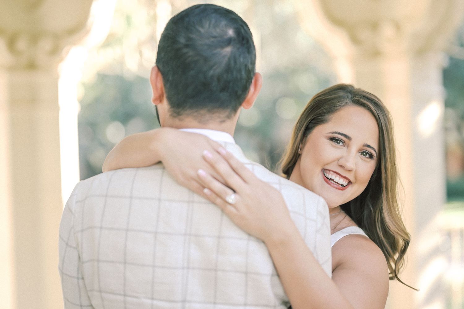 Smiling woman with long brown hair wearing an engagement ring embraces a man in a plaid shirt outdoors near stone columns.