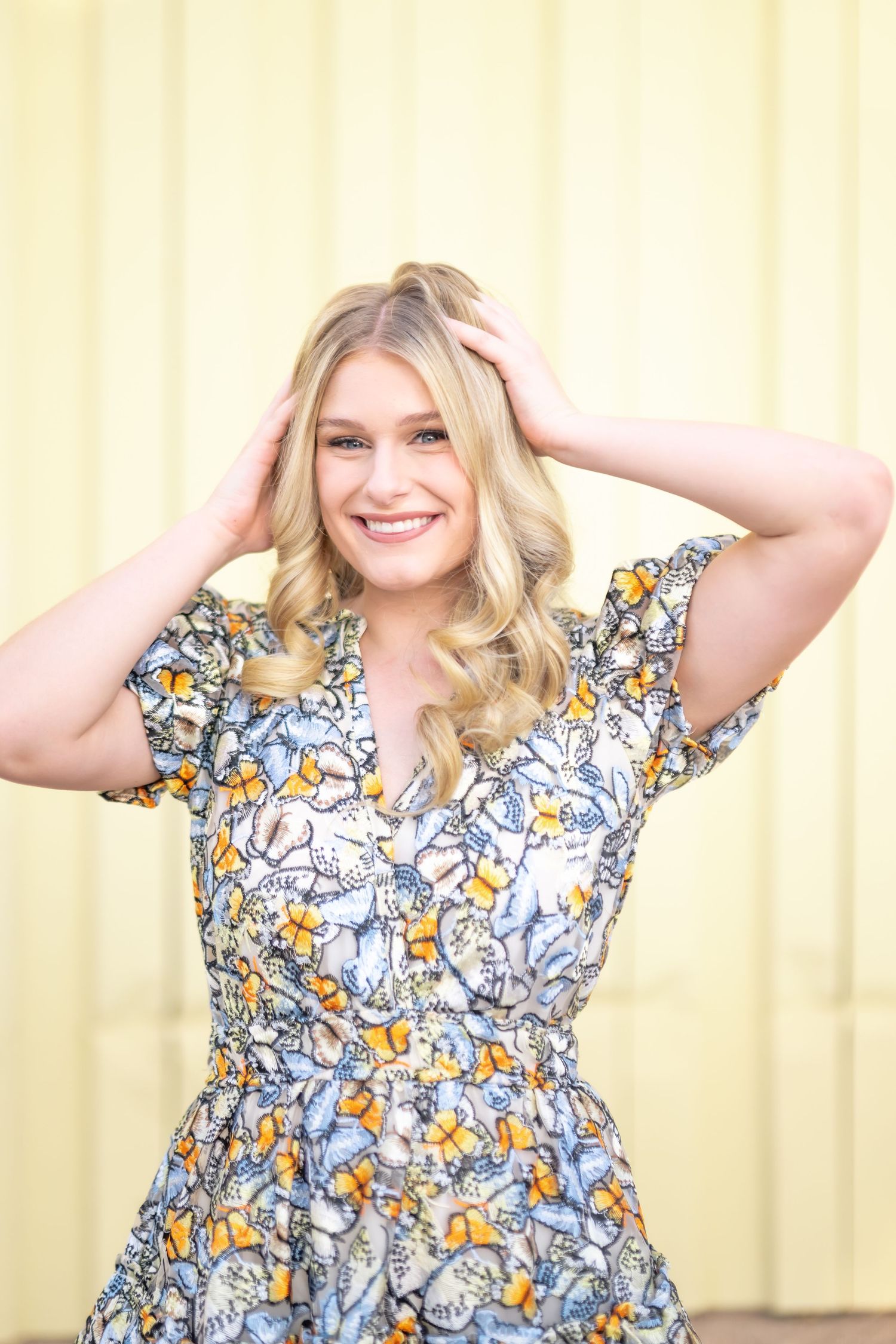 Smiling blonde woman in a floral dress with hands in hair, posing against a yellow wall.