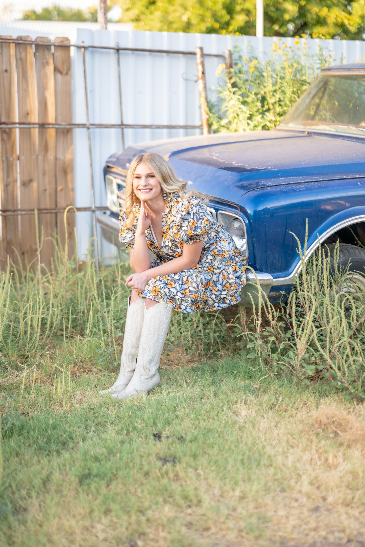 Smiling blonde woman in floral dress and white boots poses beside a vintage blue car in a grassy yard.