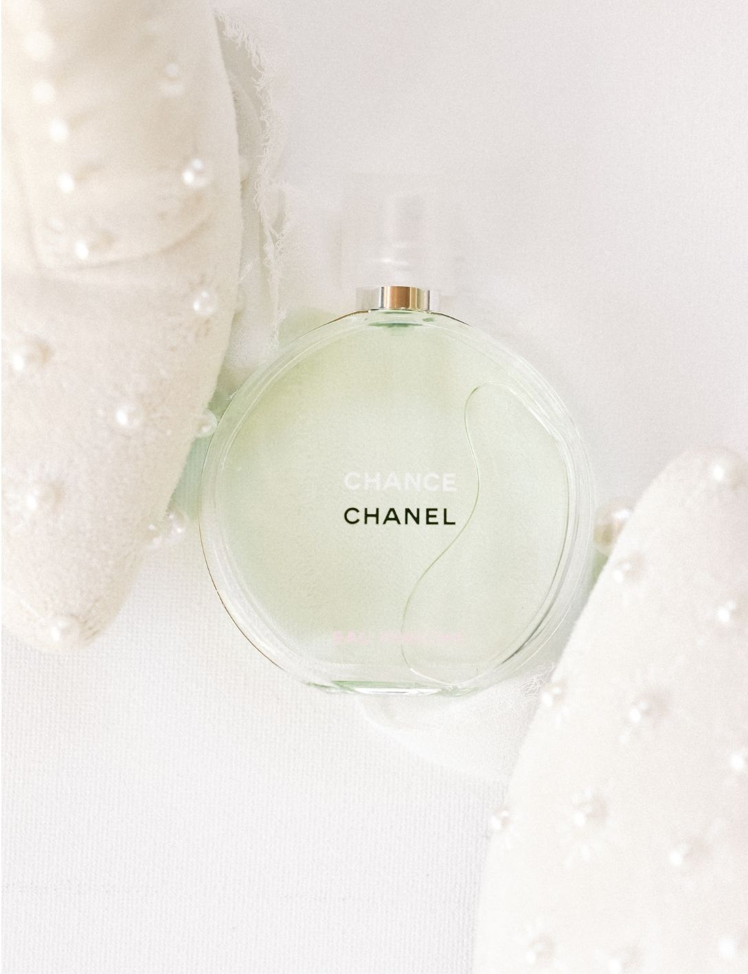 Chanel Chance Eau Fraiche perfume bottle in pale green glass lying on a white surface with pearl-adorned fabric.