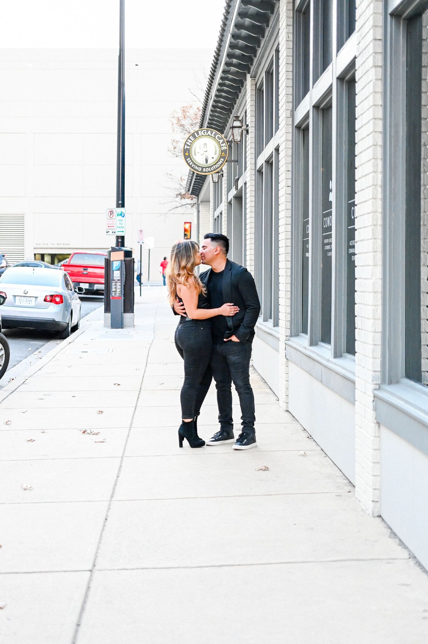 Romantic couple in black outfits sharing a close moment on a city sidewalk near a vintage street clock.